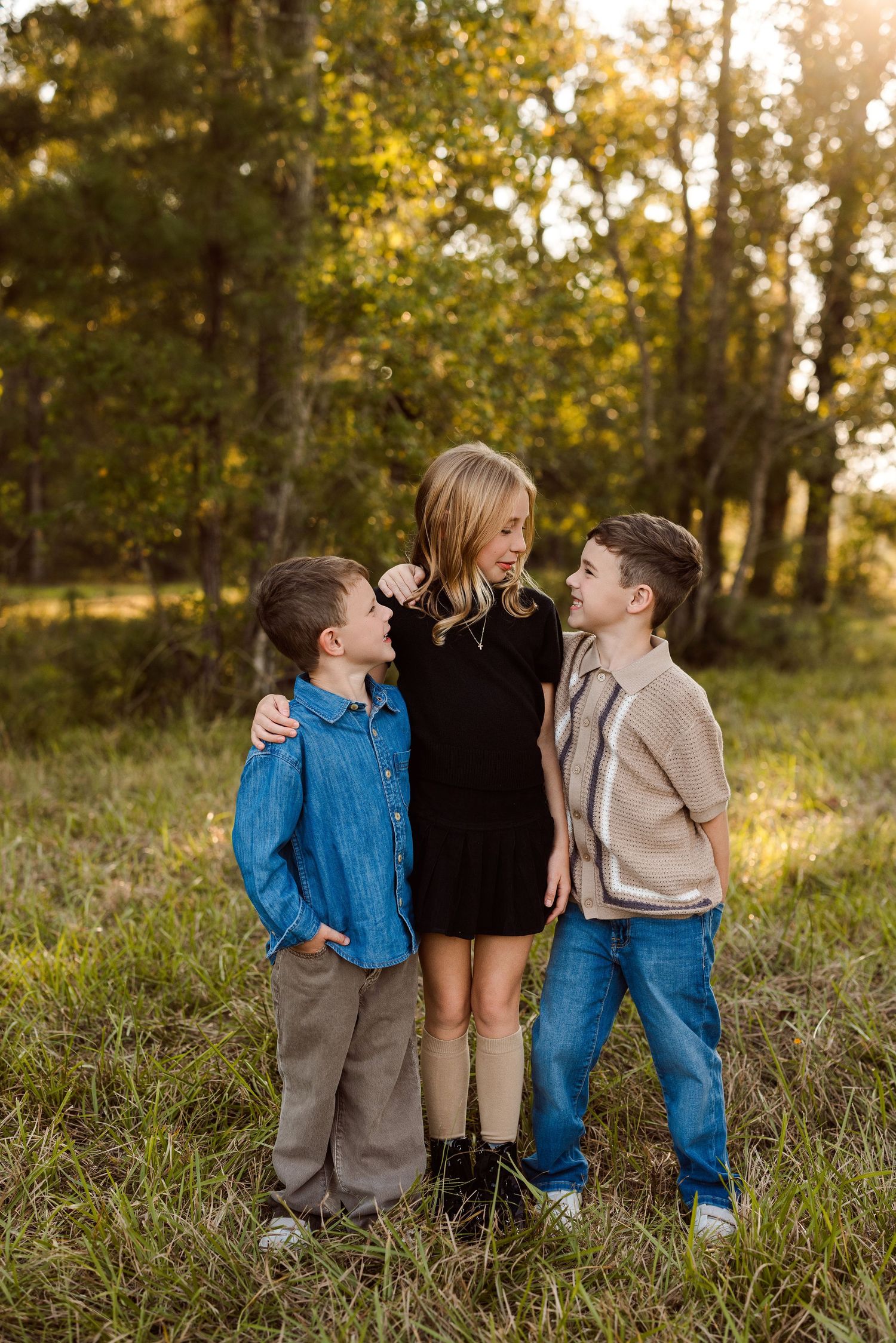 Three smiling children with arms around each other stand in a sunlit grassy field with tall trees in the background.