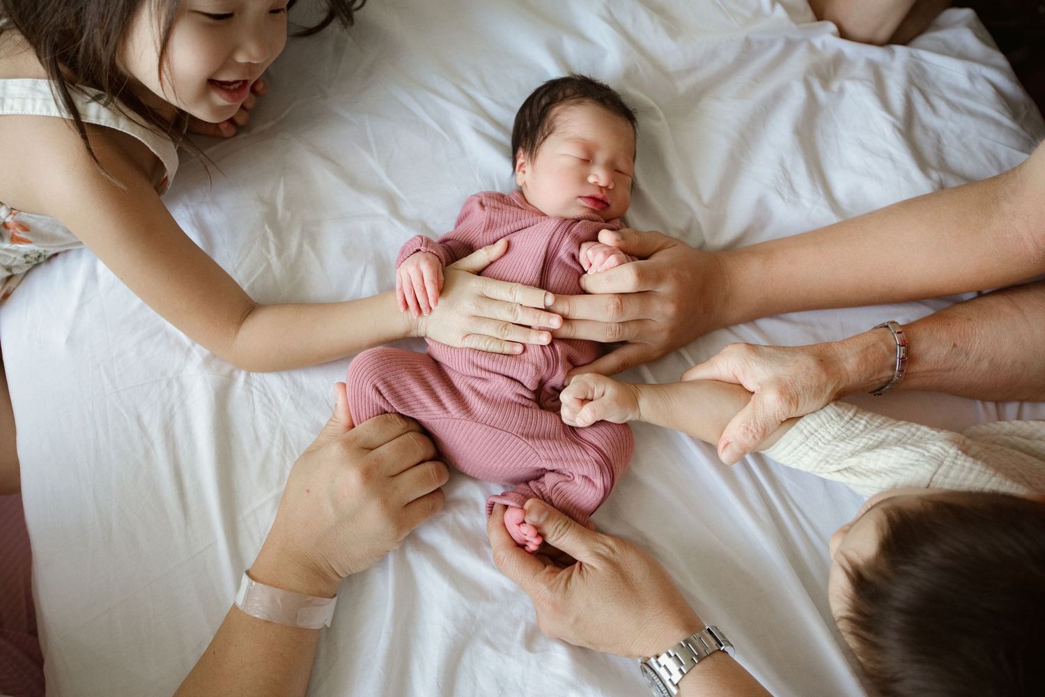 A sleeping newborn baby in pink clothing being gently held by multiple hands.