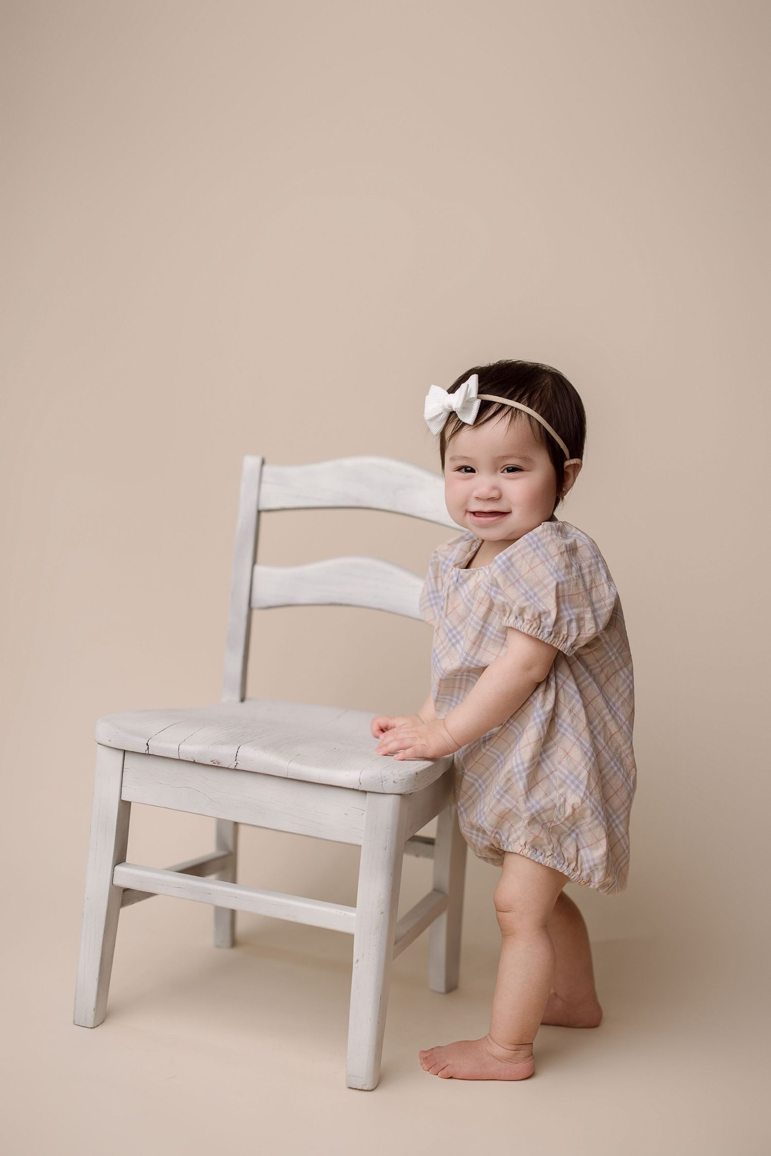 Baby smiling and playing with a small white wooden bench against a beige backdrop.
