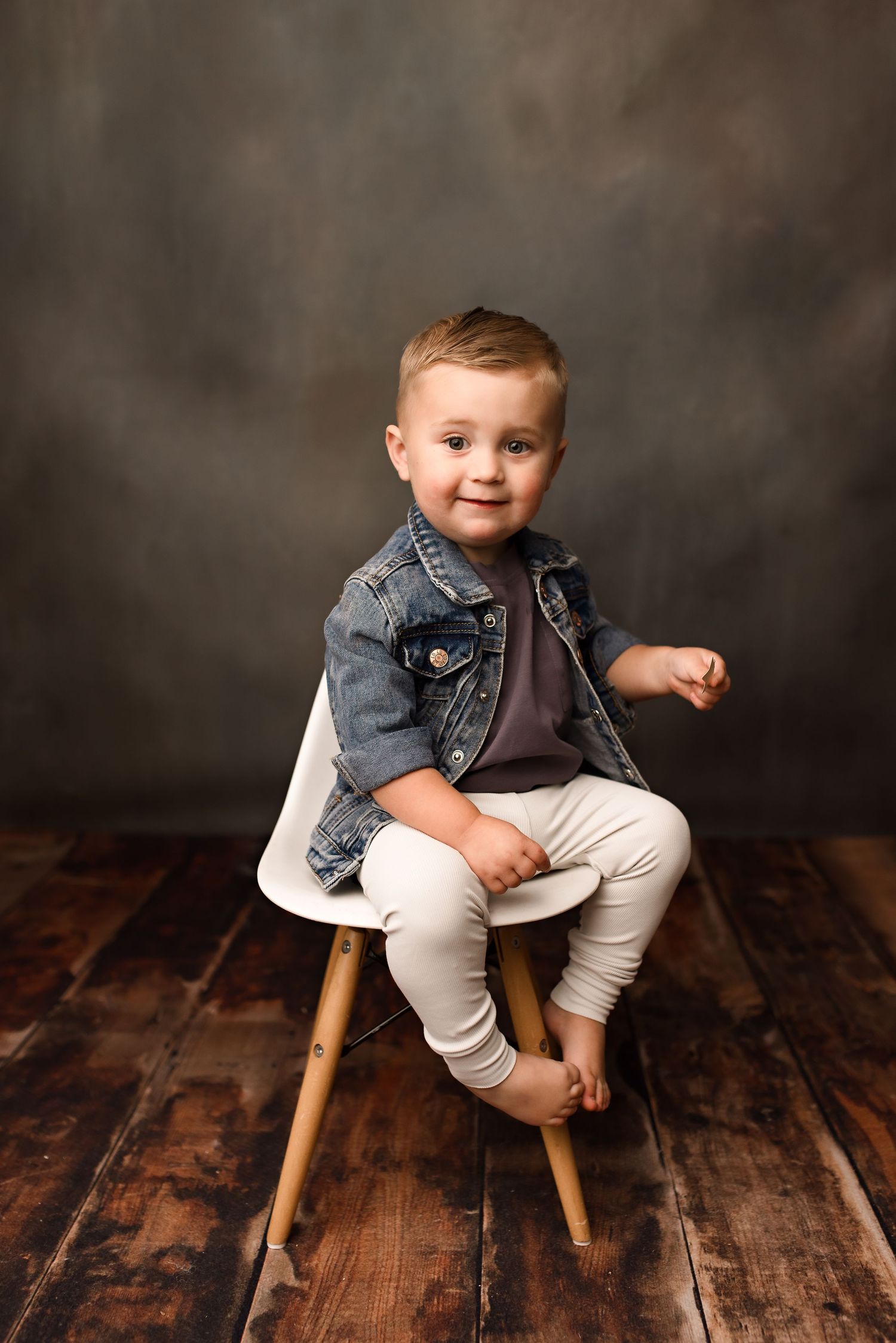 Child sitting on wooden stool wearing denim jacket during rustic studio portrait session.