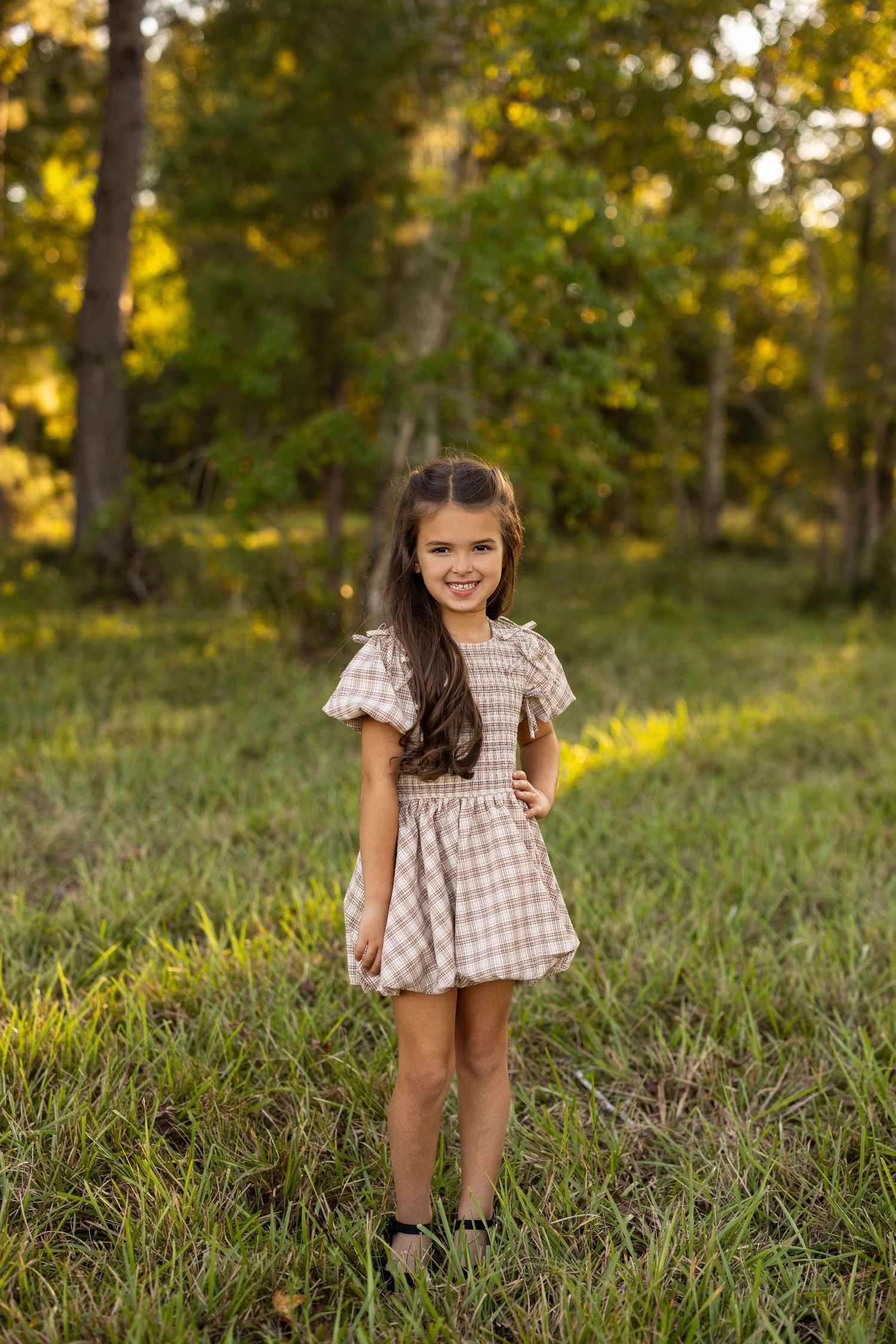 A child in a checkered dress stands in a sunlit forest clearing during golden hour.