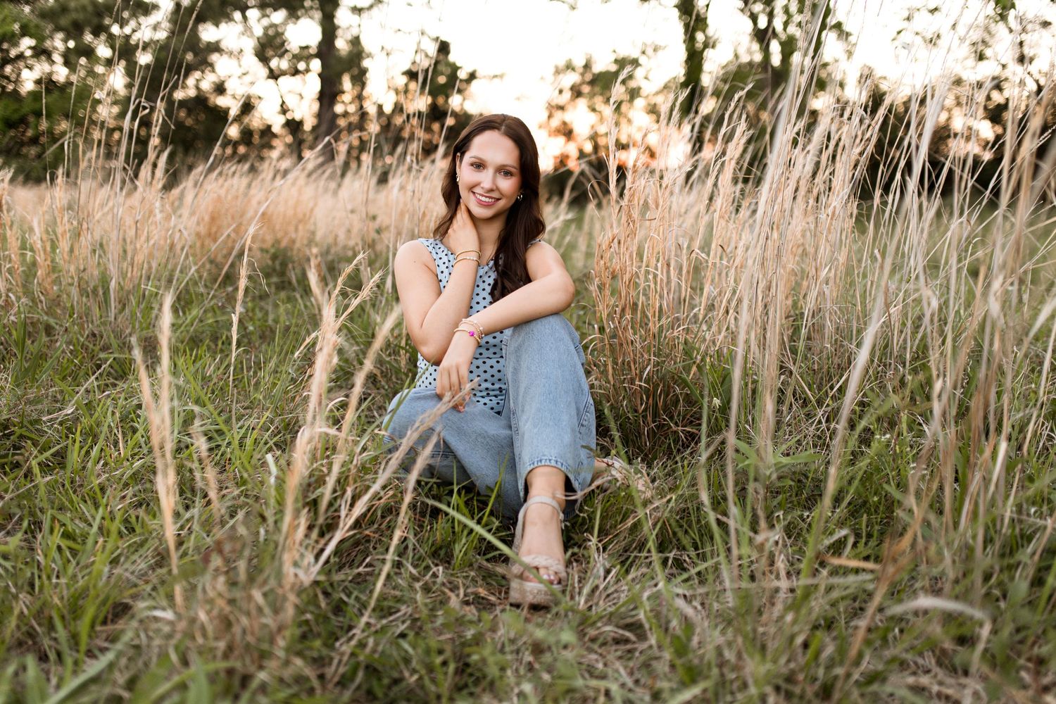 Young woman in blue outfit sitting gracefully in tall golden grass field during golden hour sunset.