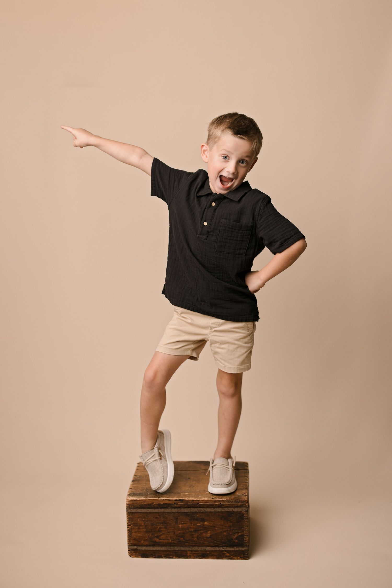 Energetic child in black polo and khaki shorts stands on wooden box while pointing and making excited expression against beige backdrop.