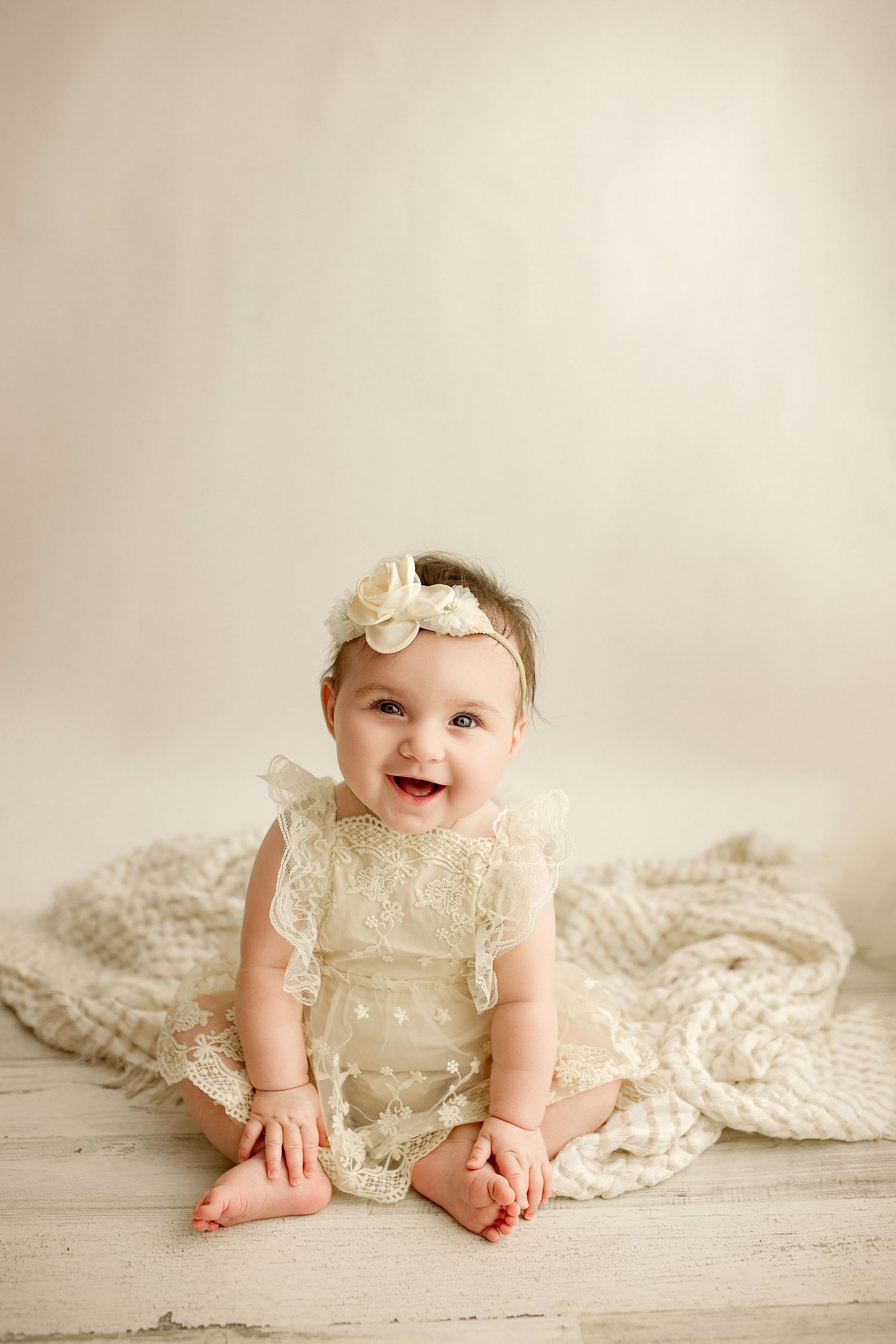 A baby wearing a cream lace dress and floral headband sits smiling on a light-colored floor with a knit blanket.