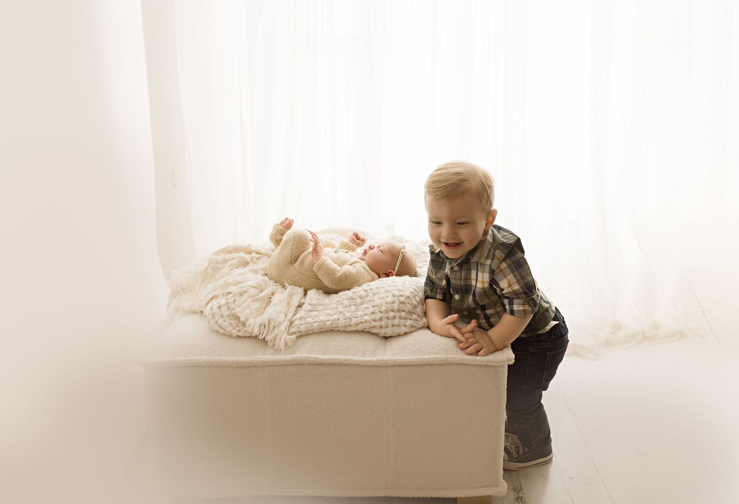 Young child in plaid shirt leans over newborn sibling on white surface.