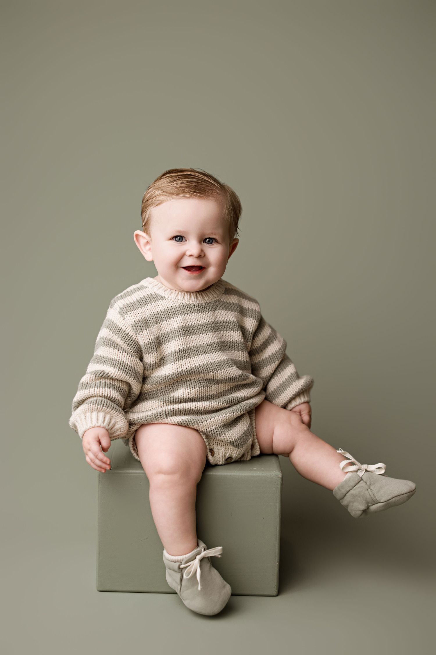 A happy baby wearing a striped knit sweater and booties sits on a gray cube against a neutral background.