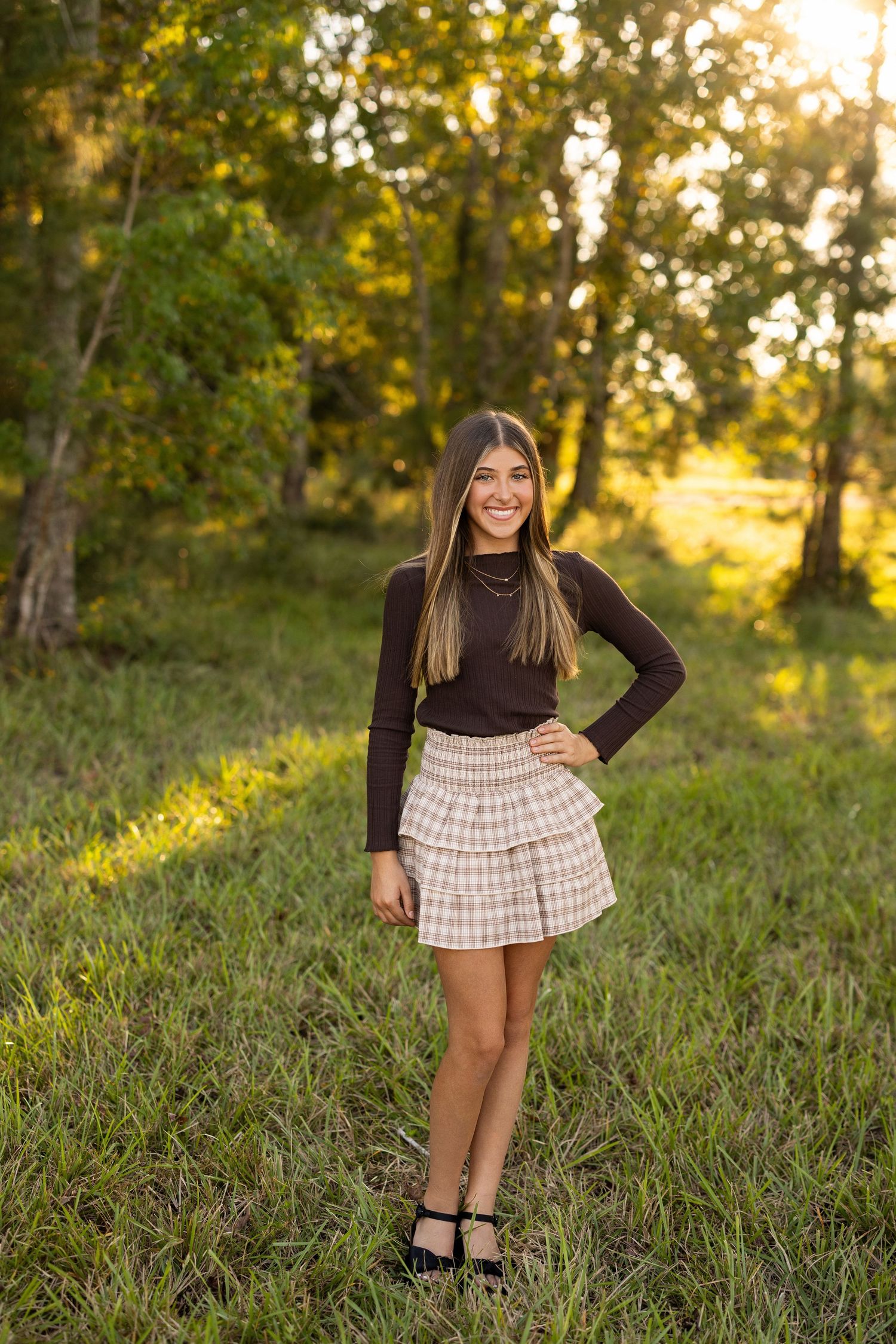 A young person in a brown top and plaid skirt poses in a sunny outdoor setting with trees.