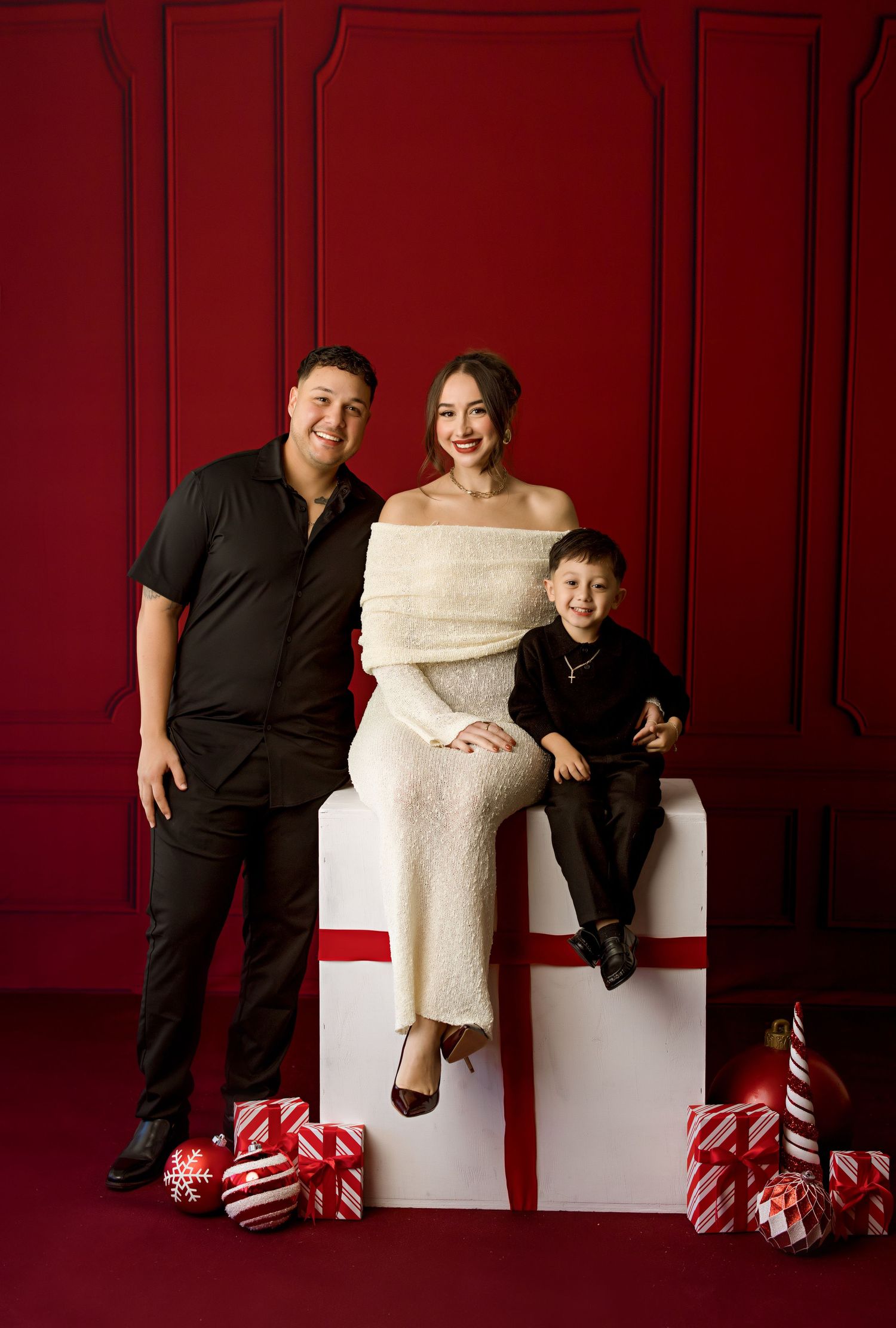 A family poses on a white gift box prop against a festive red wall with ornaments.