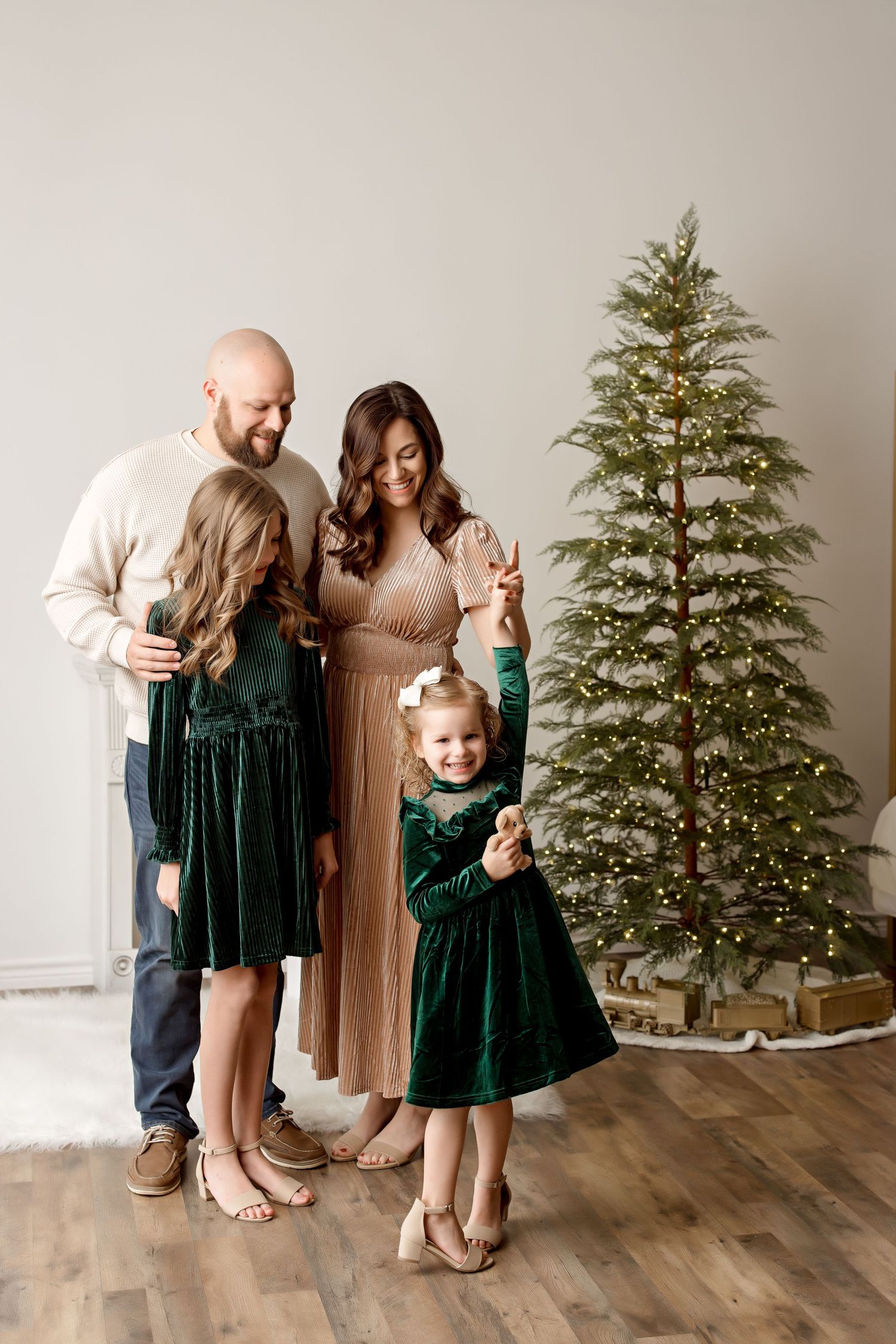 A family in elegant winter attire poses beside a decorated Christmas tree in a minimal setting.