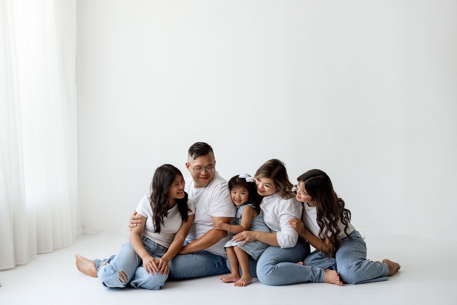 A family wearing matching denim and white shirts lies in a row on the floor of a bright minimalist studio space.