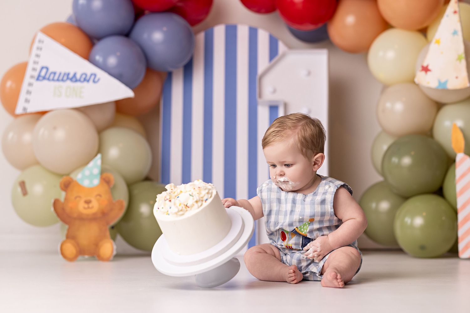 Baby in white outfit sits on floor surrounded by pastel balloons and birthday decorations for first birthday celebration.
