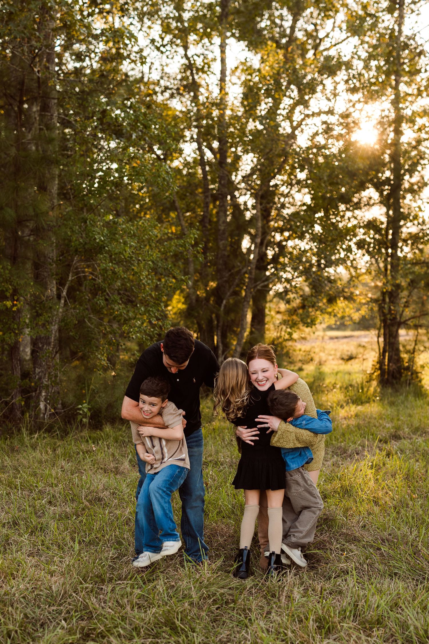 Family of five laughing and hugging in a grassy field surrounded by trees at golden hour, with warm sunlight filtering through the branches.