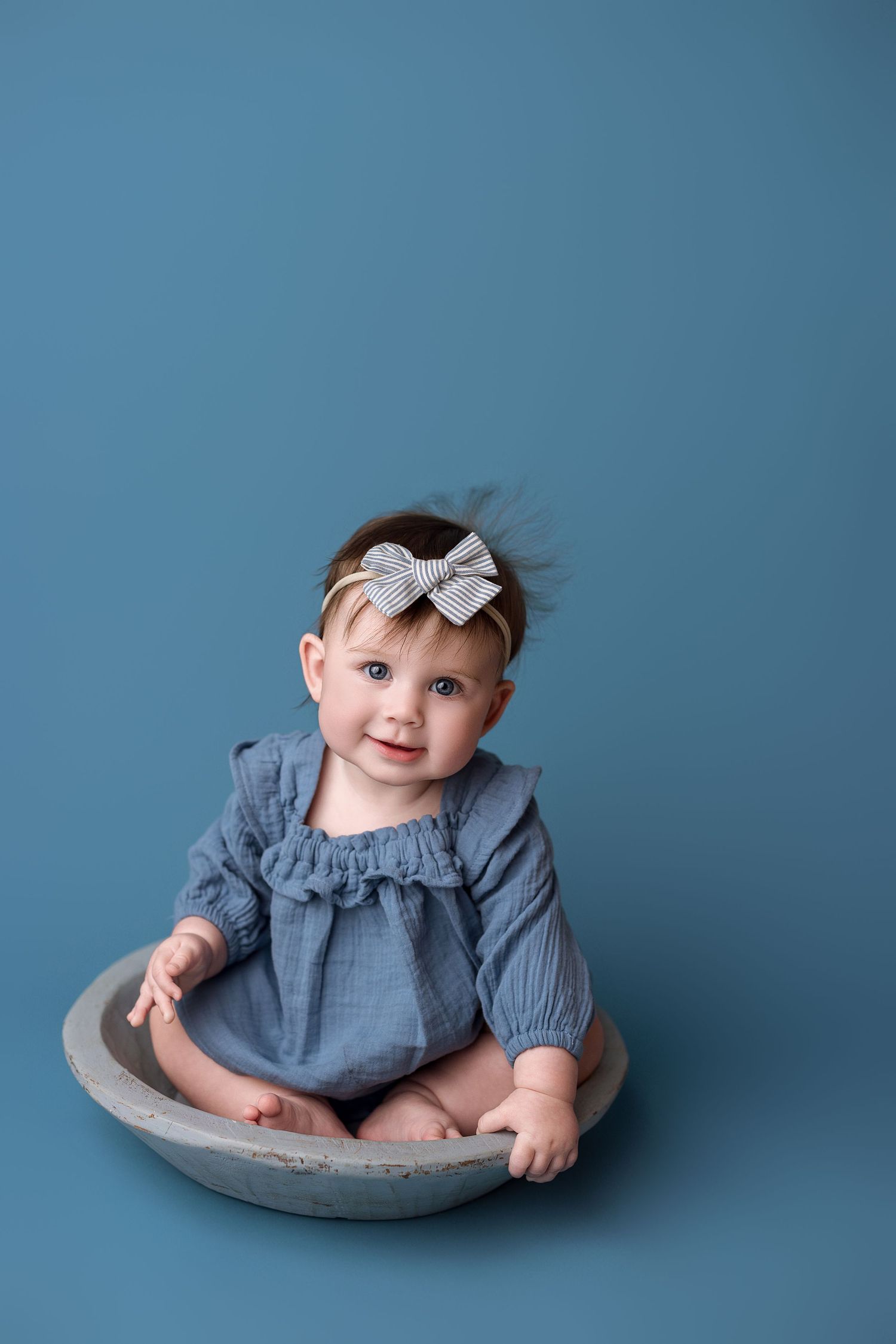 Baby wearing blue dress and bow headband sits in gray bowl against blue background.