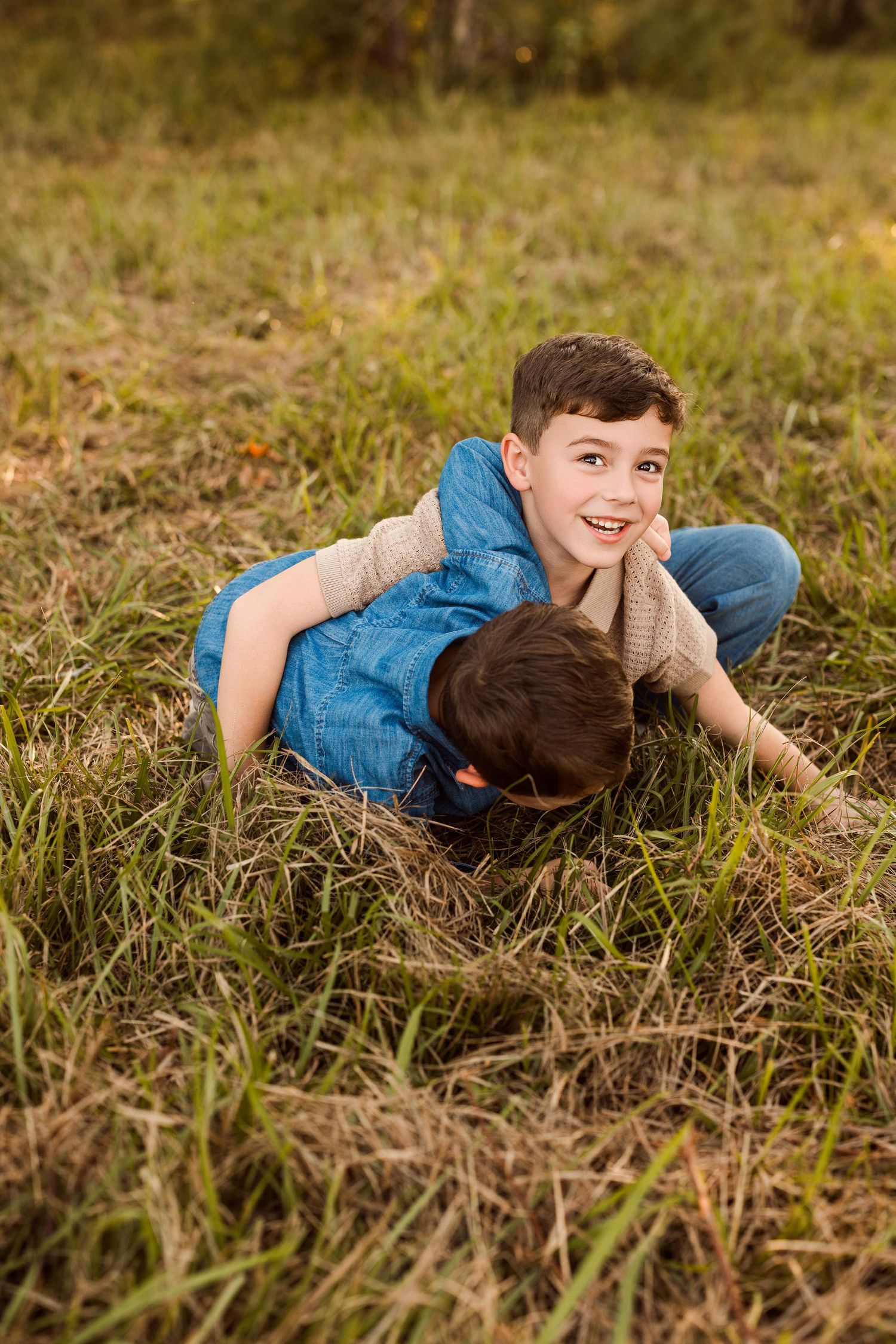 Two smiling boys in denim vests playfully wrestle on a grassy field during golden hour, one laughing at the camera.