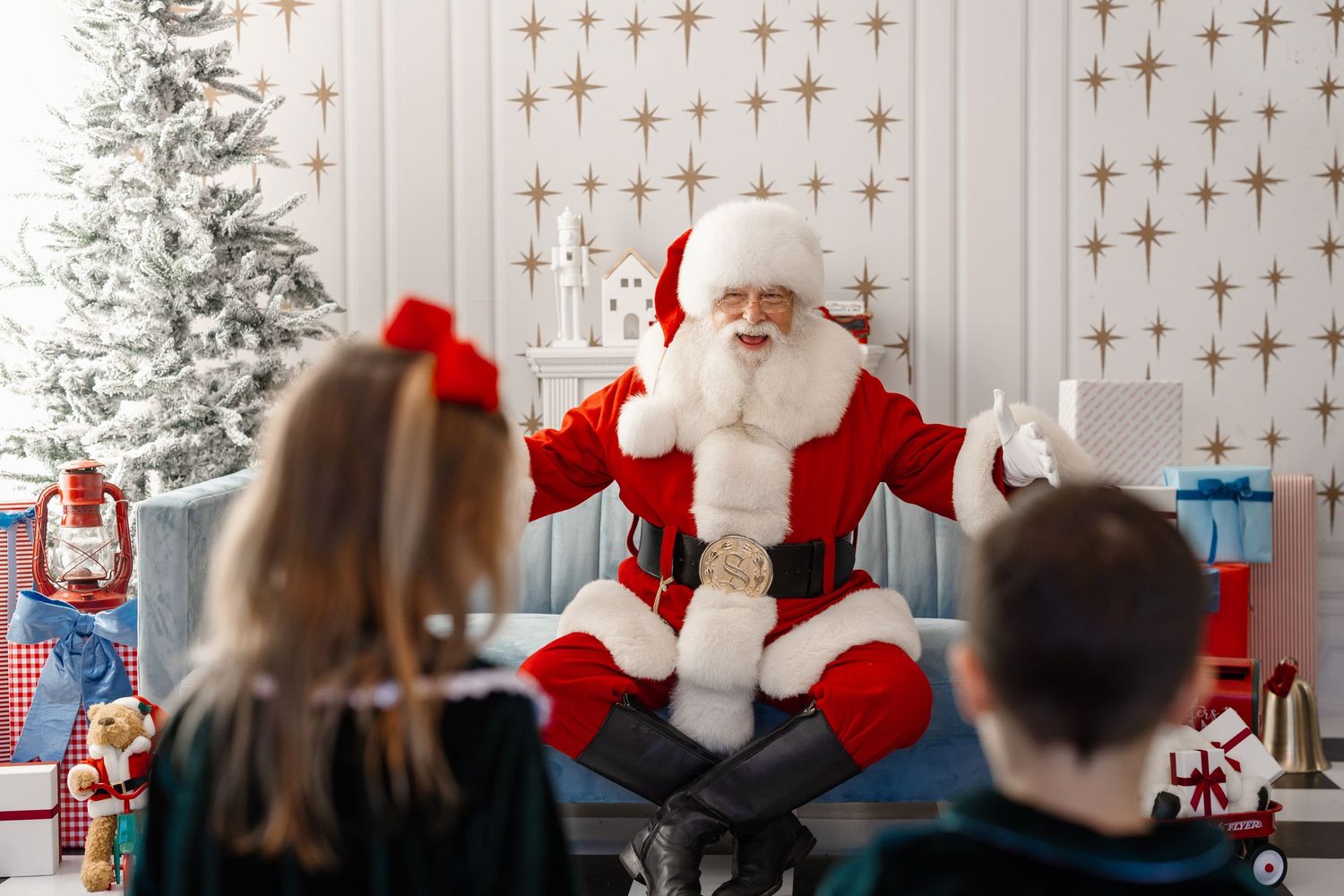 Children visit with Santa Claus in front of a decorated Christmas tree and twinkling light backdrop during holiday season.