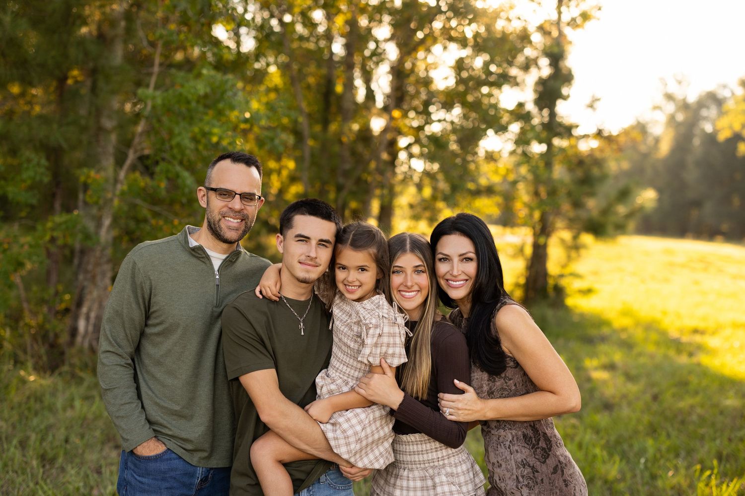 A loving family poses together outdoors in a sunlit field with trees during golden hour.