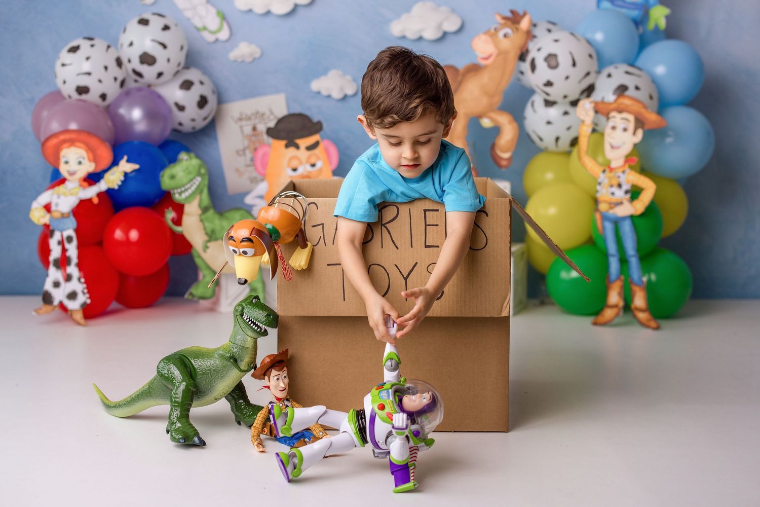 Toy Story character figurines arranged in a playful scene with cardboard boxes and colorful balloons in the background.