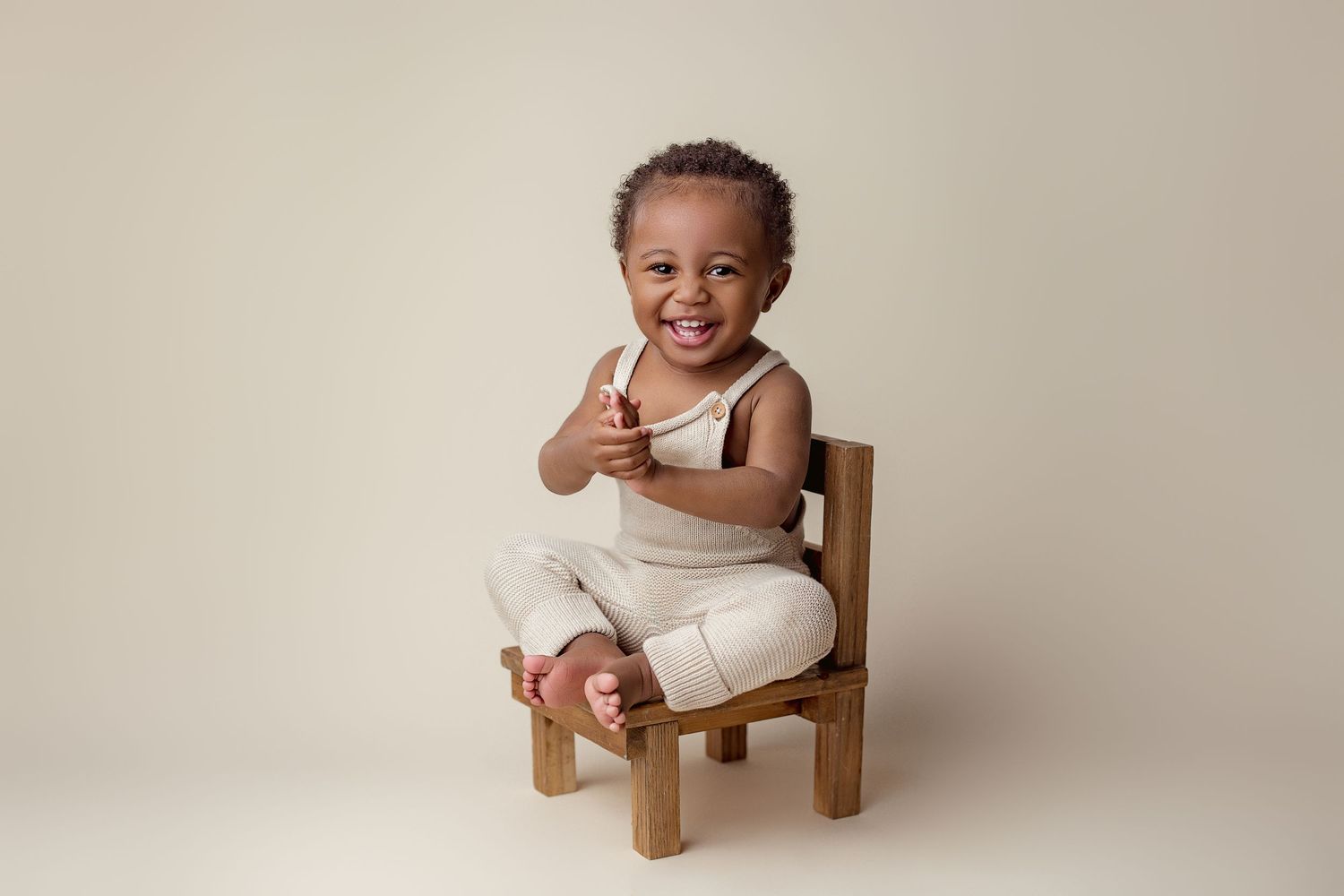 A smiling baby wearing a white onesie sits in a small wooden chair against a neutral backdrop.