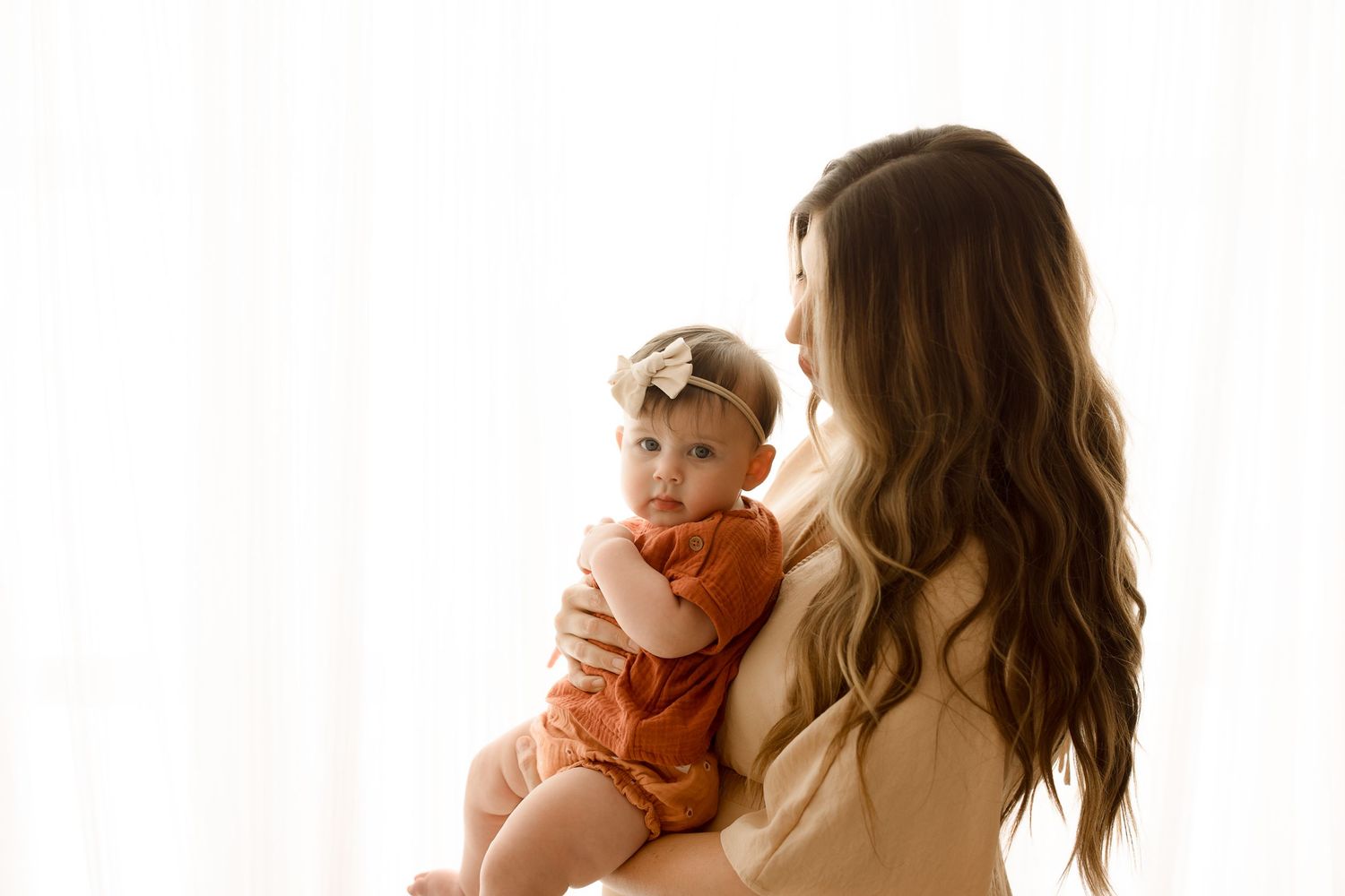 Person with long wavy hair holds baby in orange romper near window with natural light.