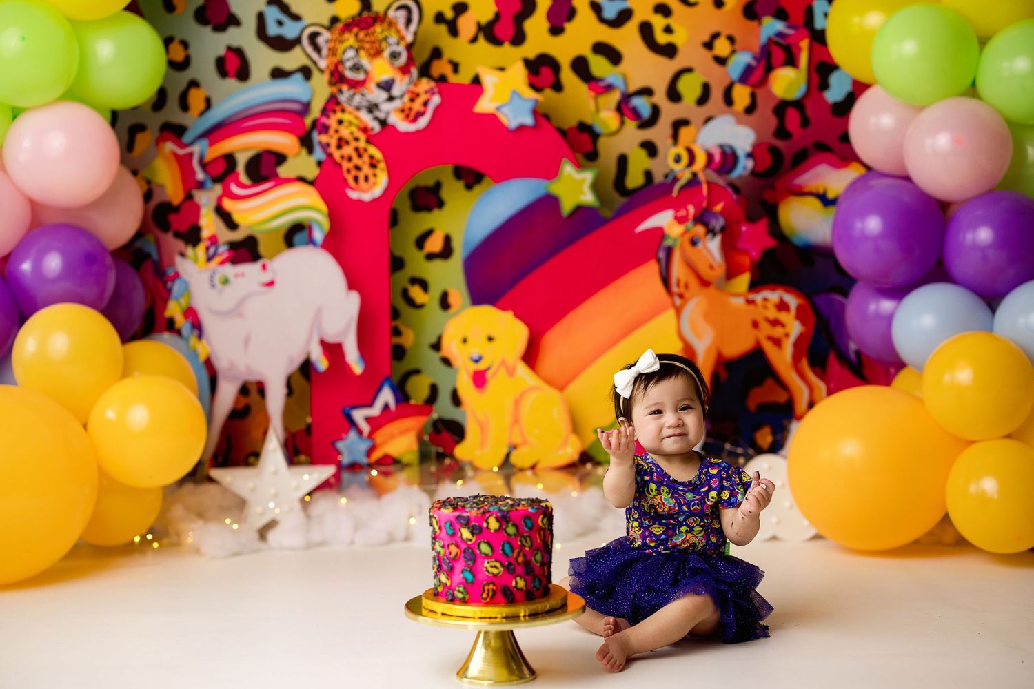 Colorful birthday party setup with leopard print backdrop, rainbow decor, balloon arches and pink cake on a stand.