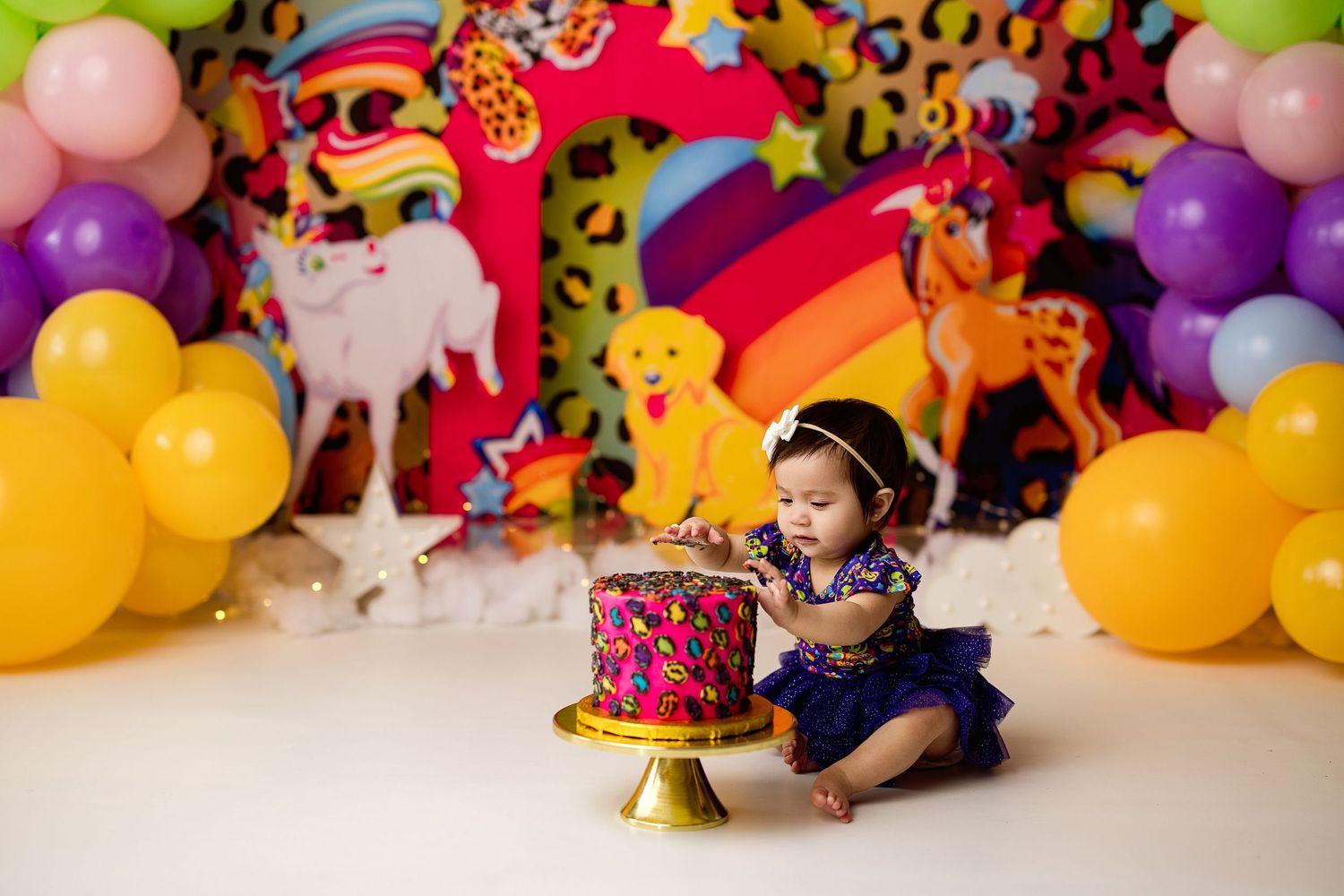 A small child sits beside a pink birthday cake with colorful rainbow and cheetah print party decorations and balloons behind.