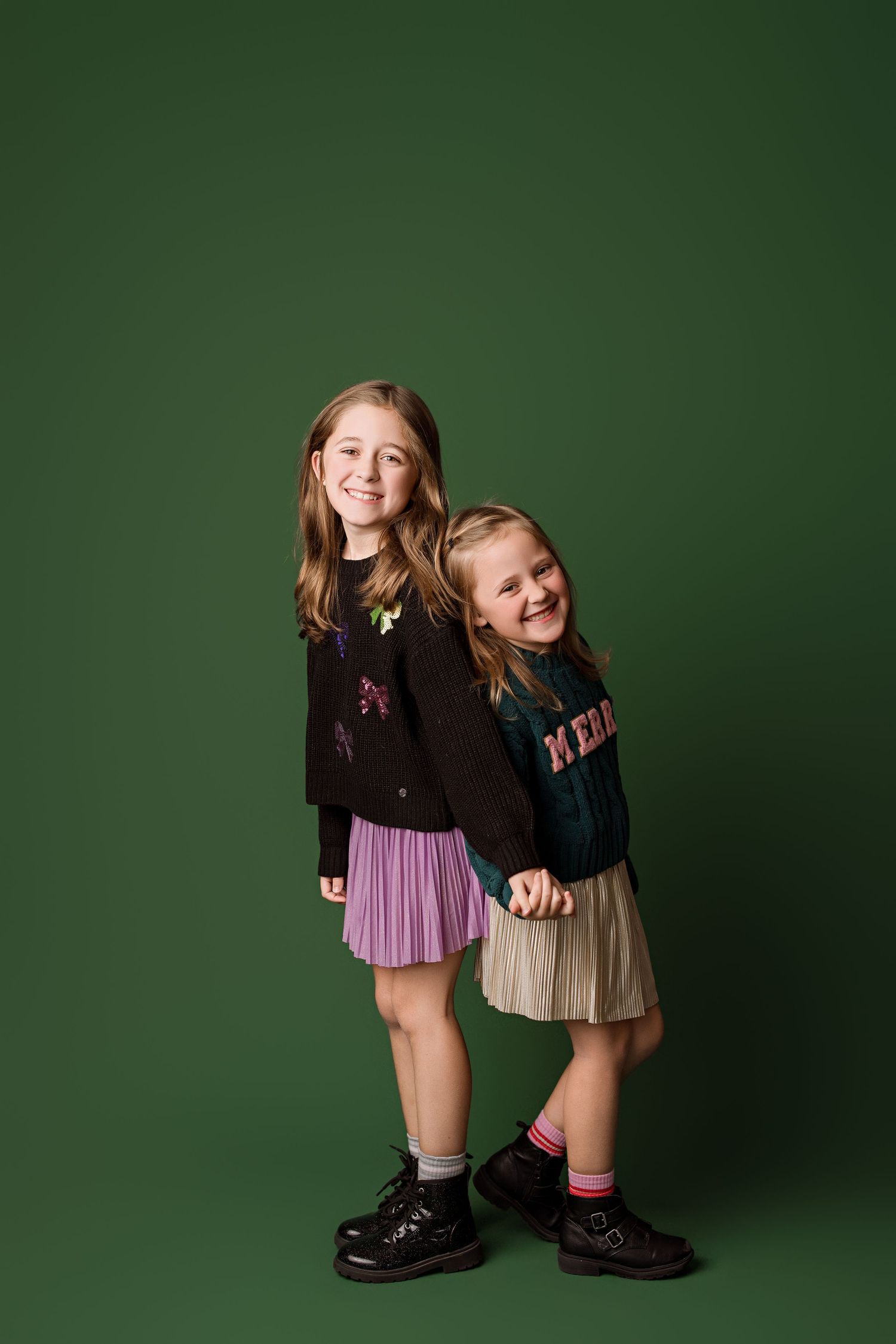 Two children wearing black sweaters and pleated skirts pose playfully against a dark green studio backdrop.