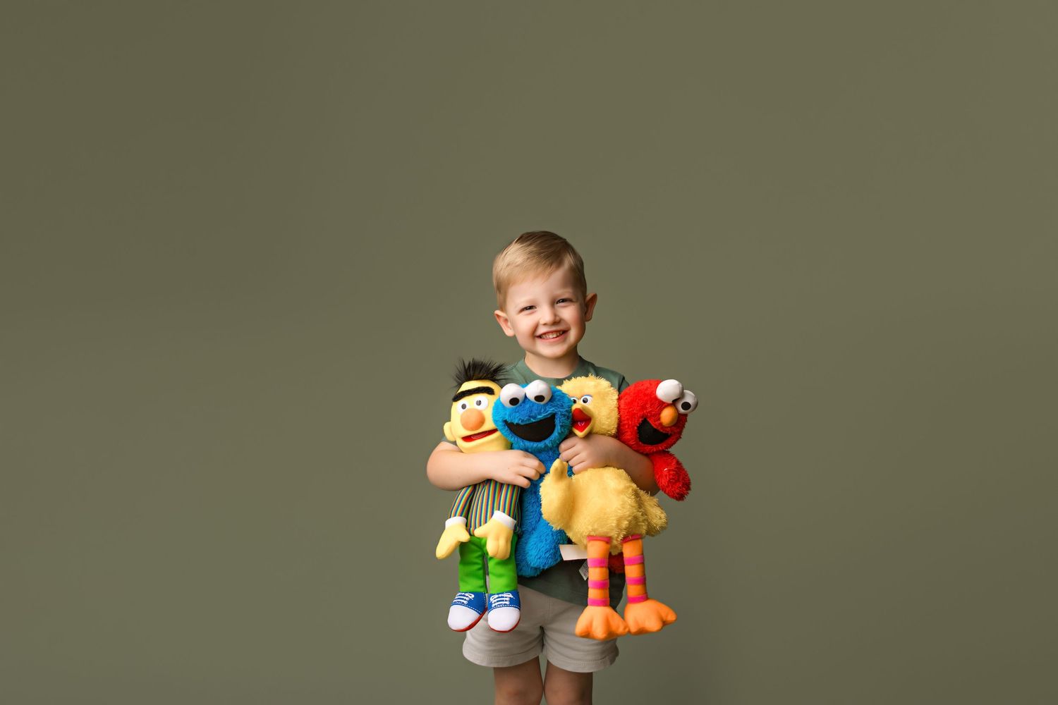 Child holding colorful stuffed animal toys against olive green backdrop.