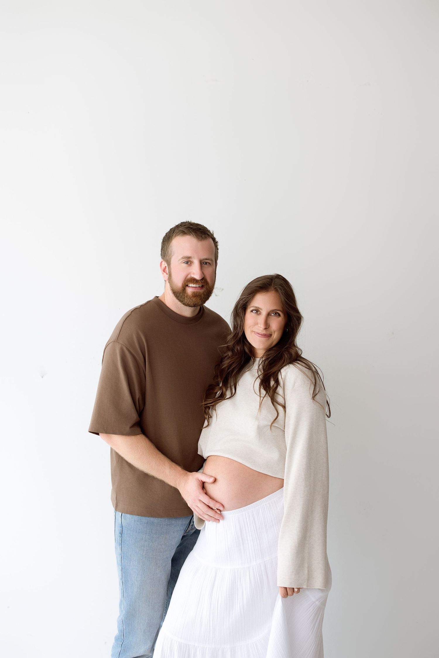 A couple poses for maternity photos in white and brown clothing against a neutral background.