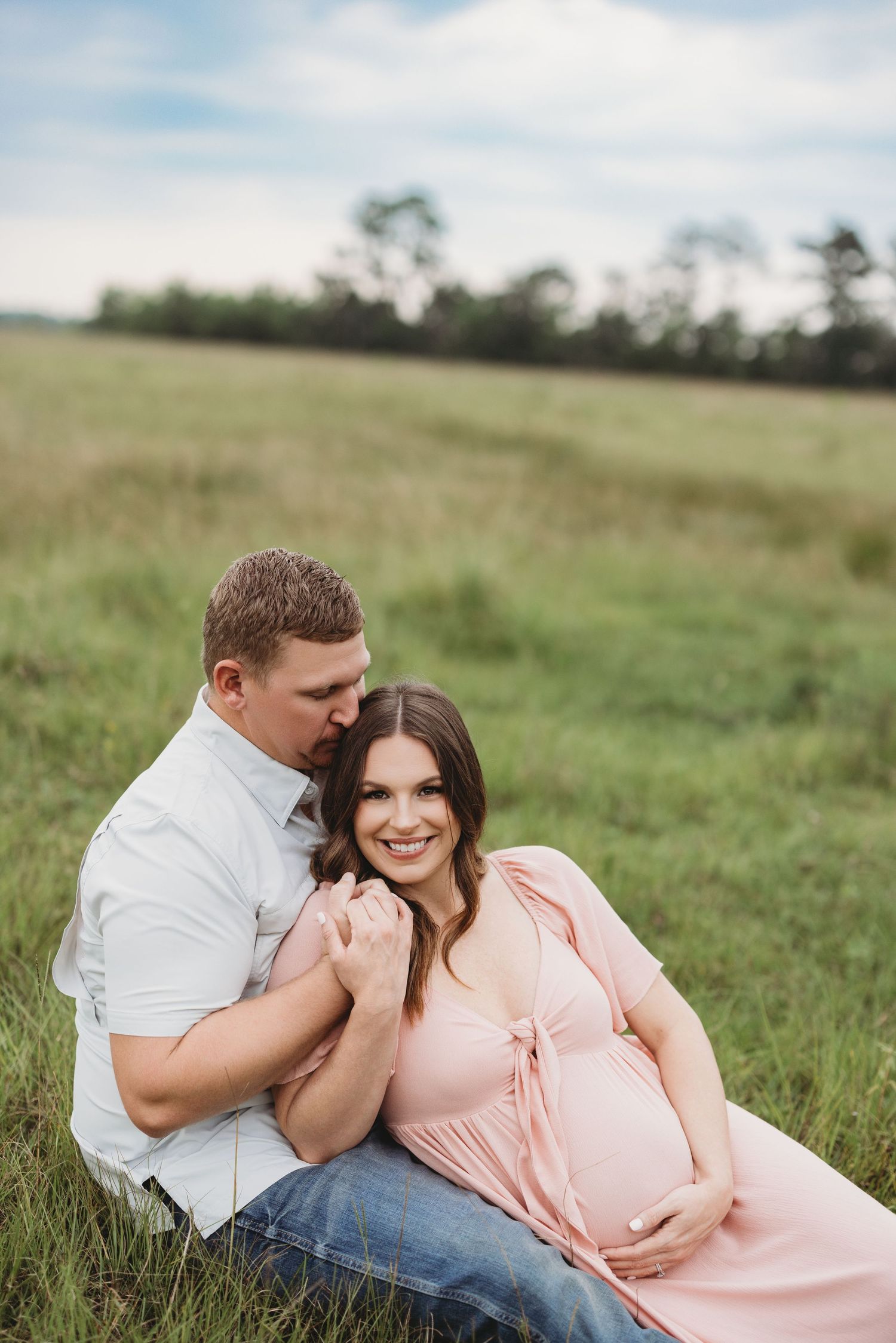 A couple shares tender moments while laying in a grassy meadow, wearing a pink dress and white shirt with Bree Elle Photography in Orange