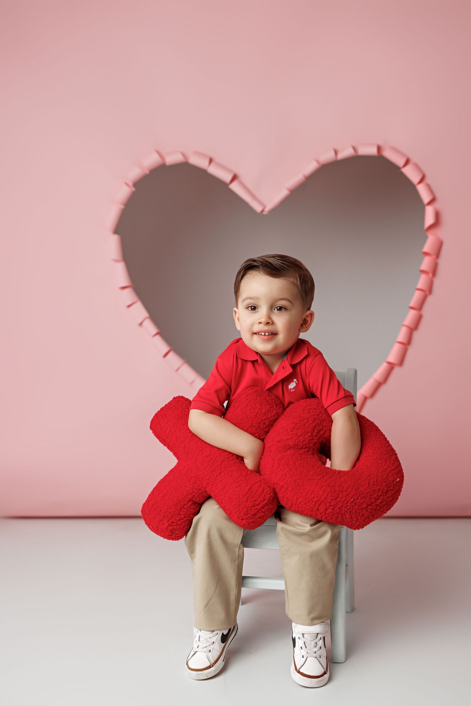 Child in red sweater posing in front of heart-shaped cutout on pink background.
