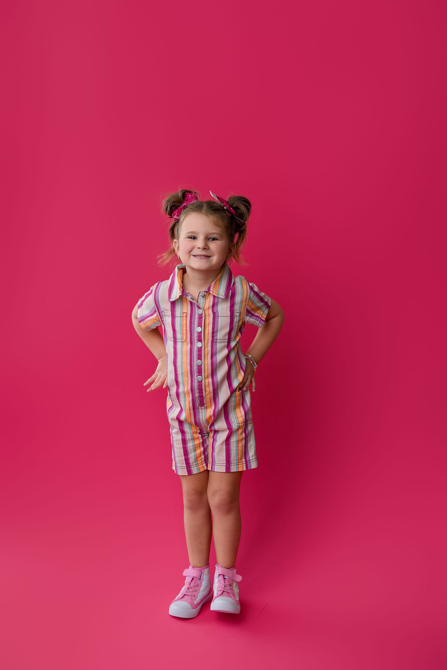 Child wearing pink striped shirt and white shoes poses playfully against vibrant pink background.