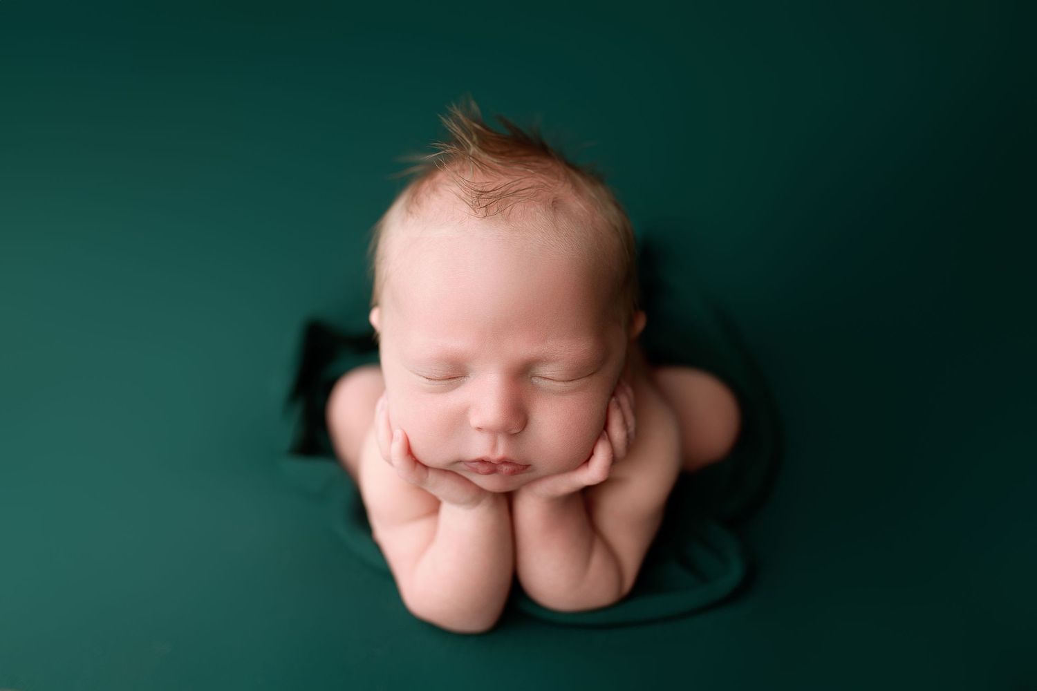 Sleeping newborn baby resting on elbows against emerald green backdrop in professional studio portrait.