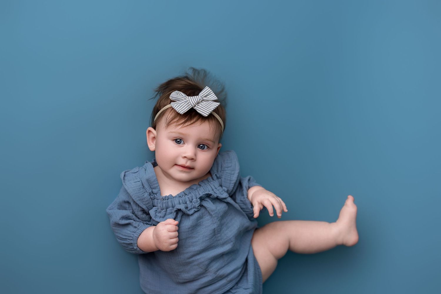 Baby wearing denim dress with white bow headband lying against blue background.