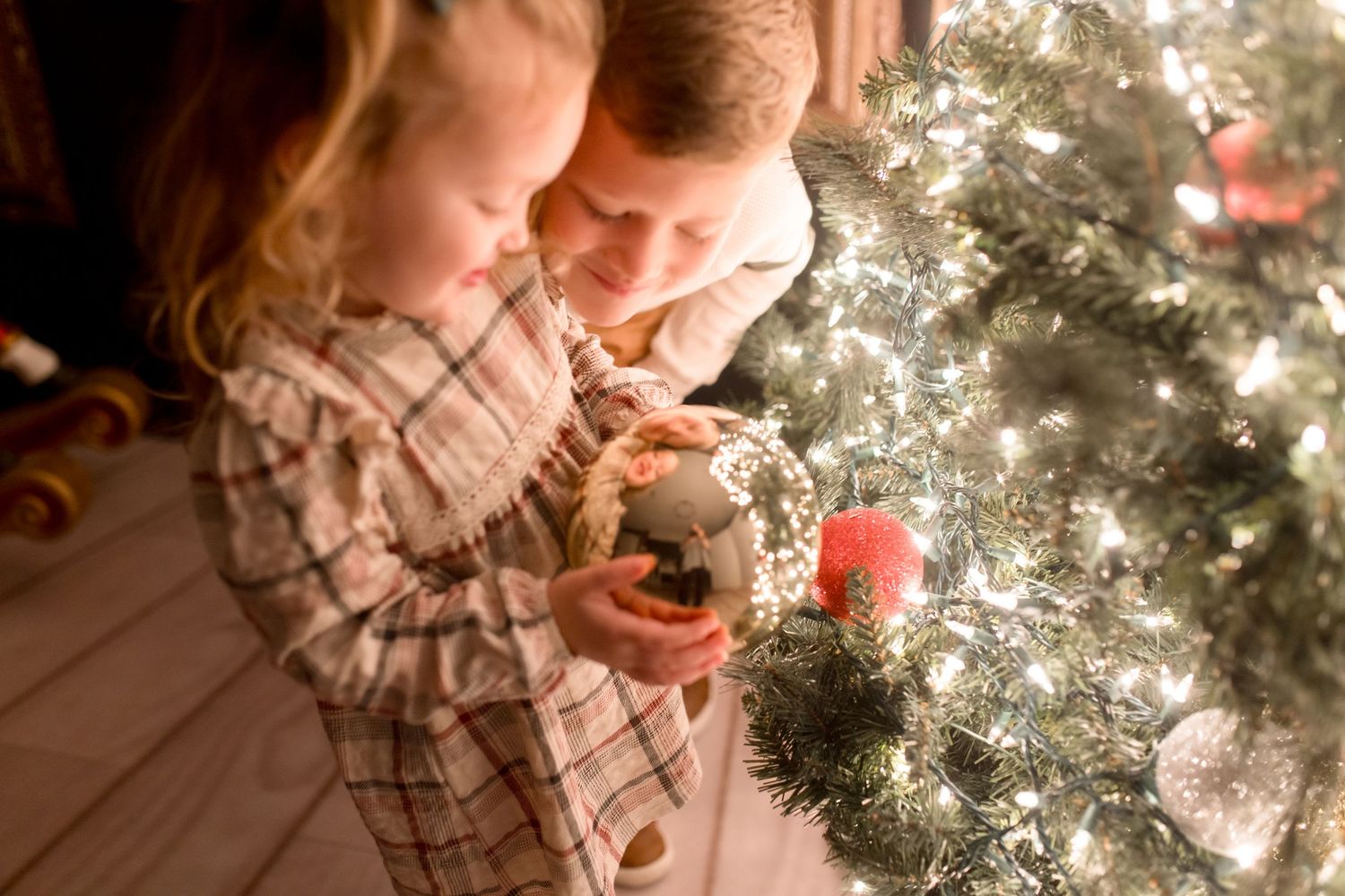 Two people lean in close together while decorating a glowing Christmas tree with ornaments and lights.