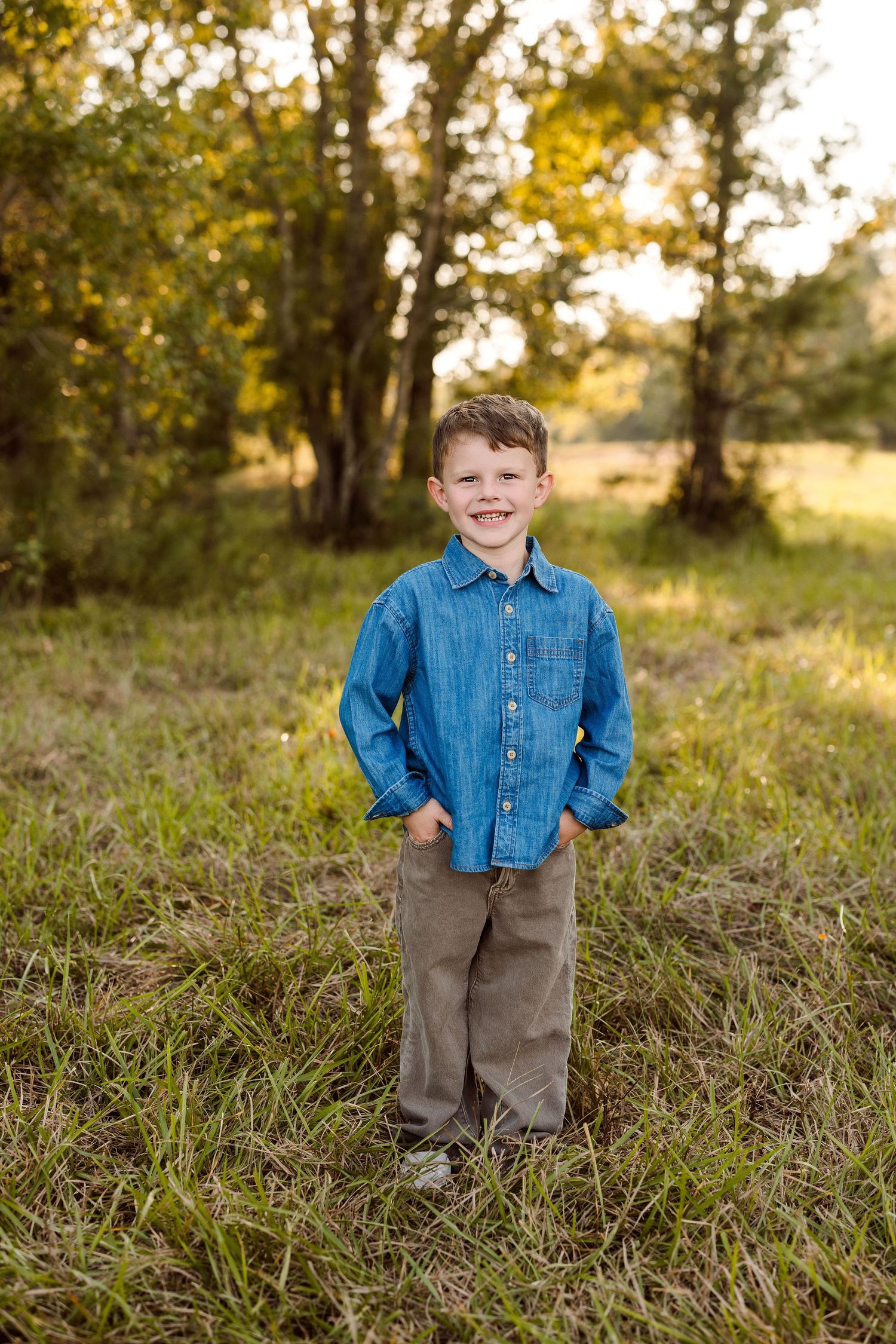 Smiling young boy in a blue denim shirt and khaki pants stands with hands in pockets on a grassy field with trees in golden sunlight.