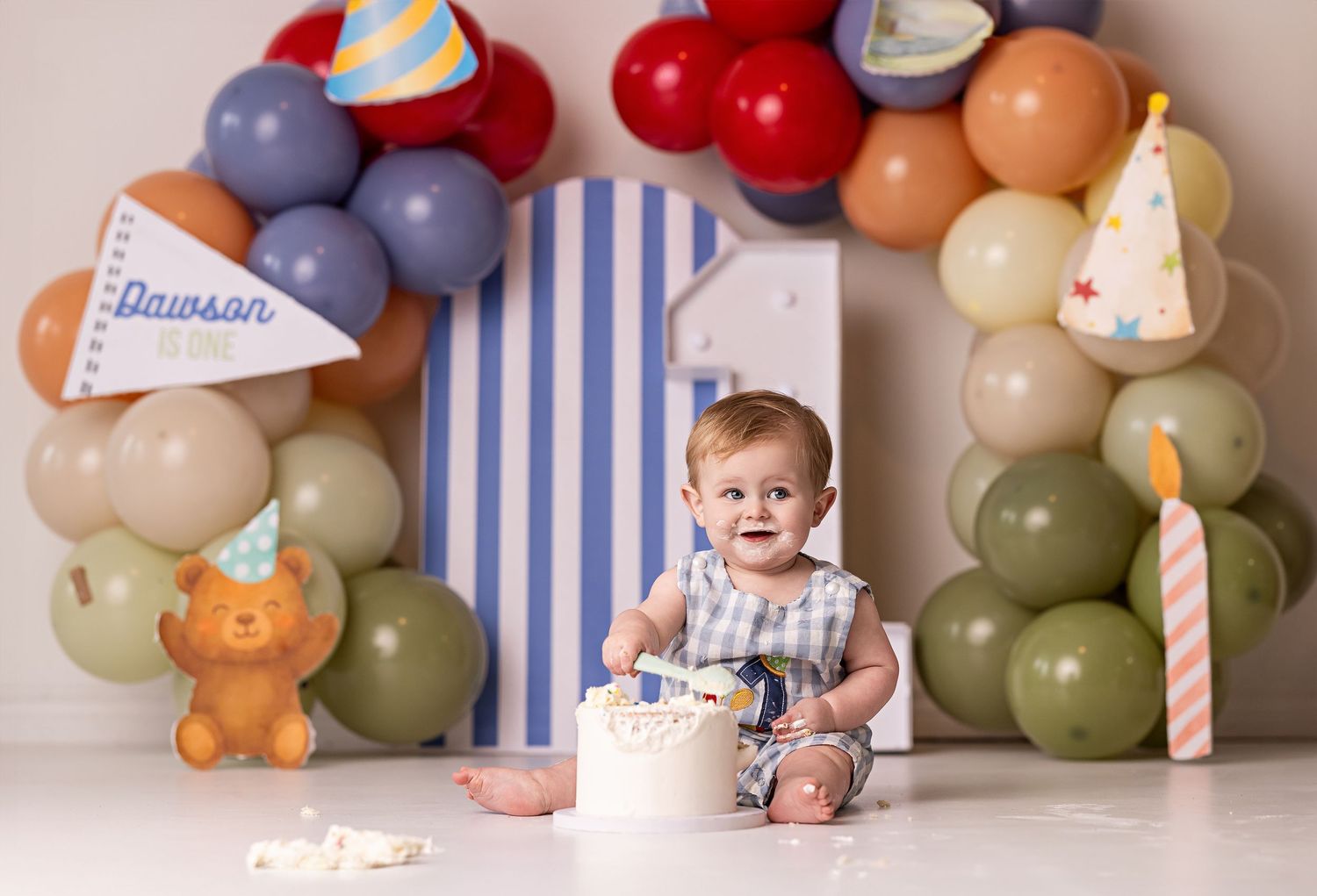 Colorful balloon arch with party hats decorates a first birthday cake smash photo session setup with blue stripes backdrop.