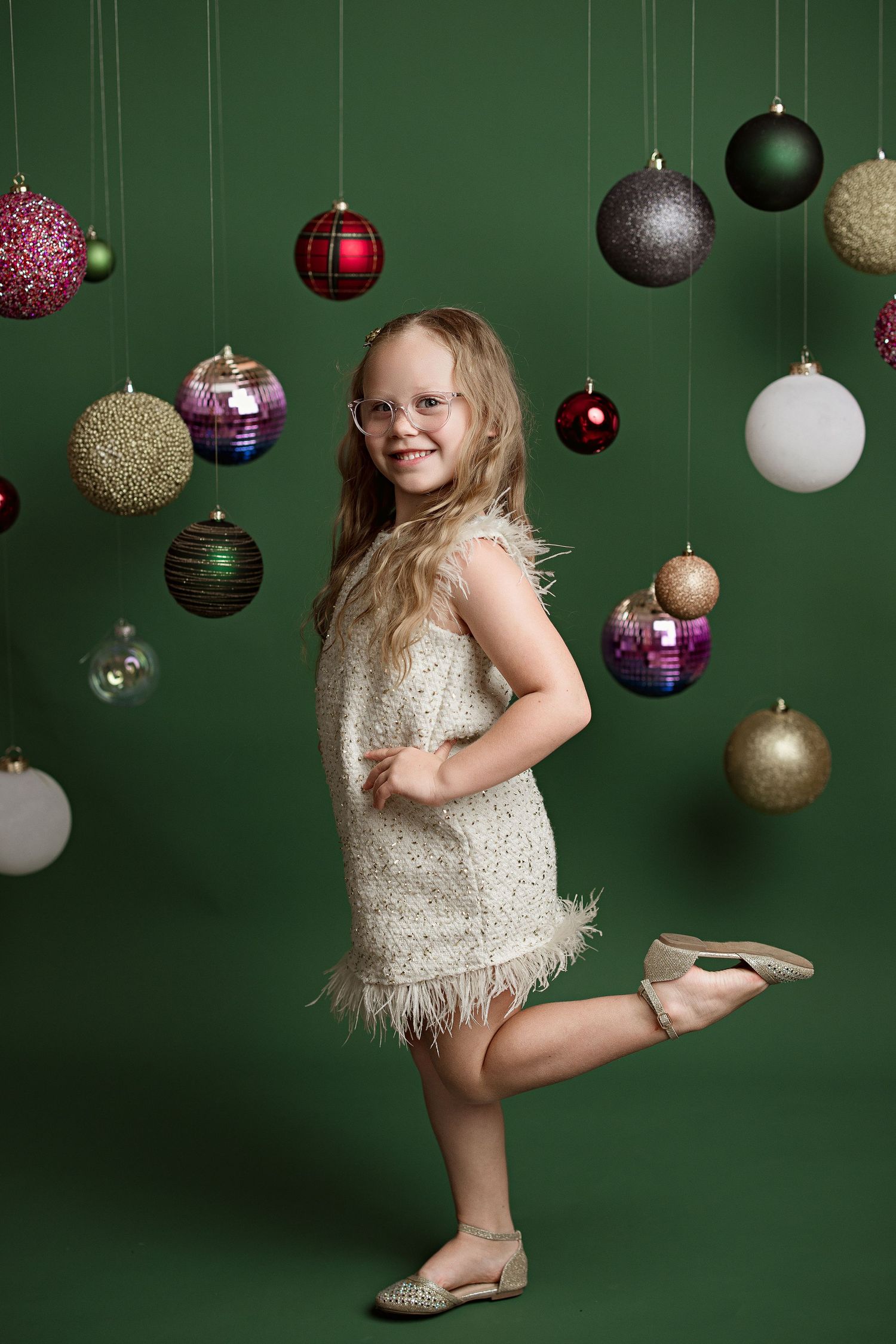 A child in a sparkly dress dances among hanging Christmas ornaments against a dark green backdrop.