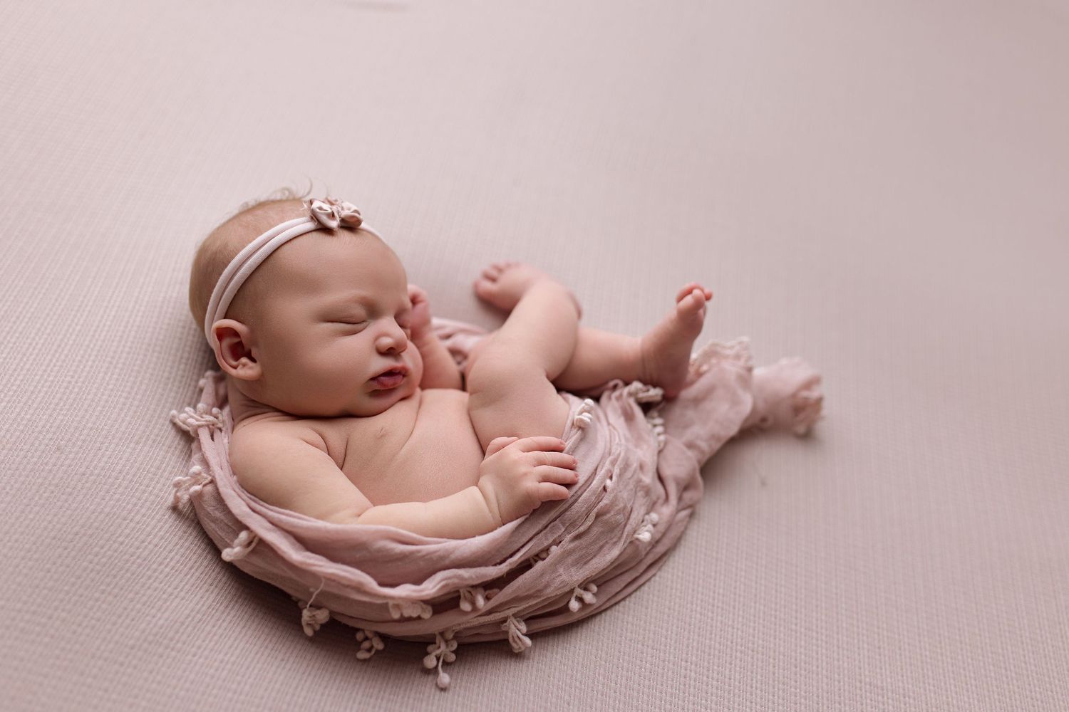 A sleeping newborn baby lies on soft pink fabric in a curled position during a professional photo session.