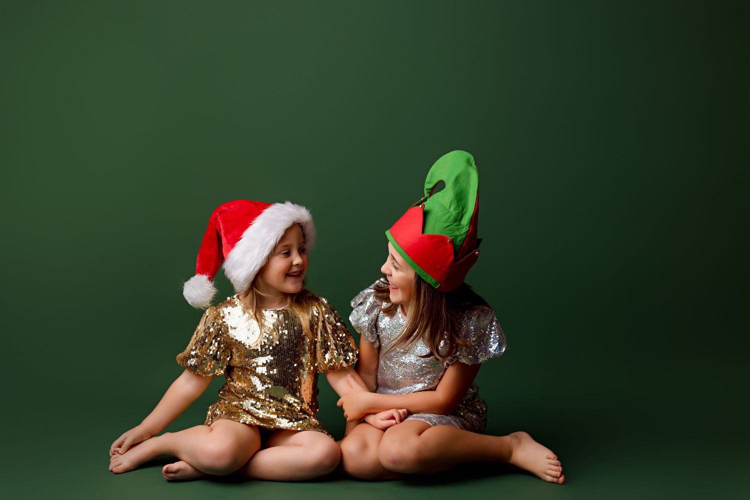 Two children in sparkly dresses wearing Christmas hats sit on floor against green background.