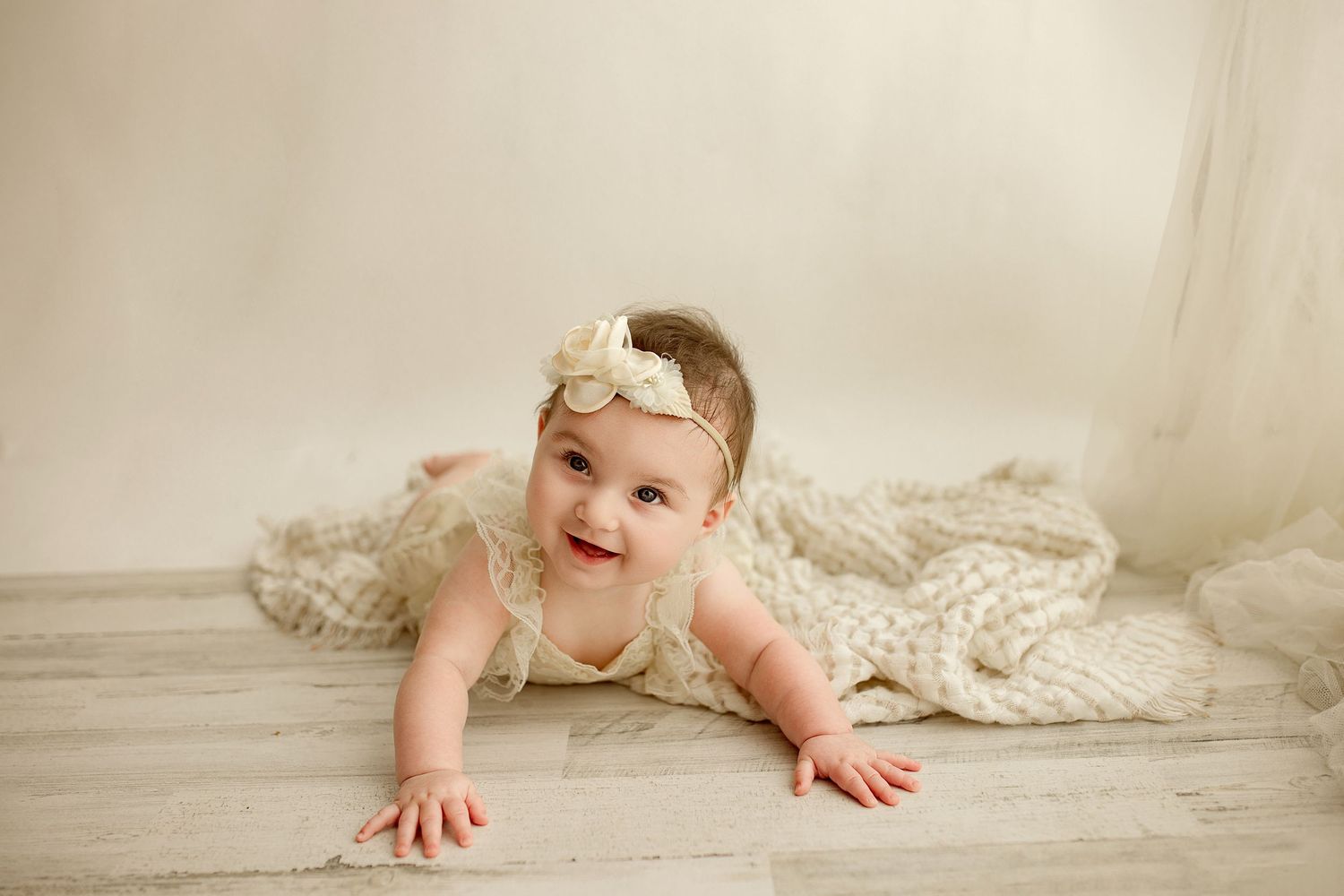 A baby in a white lace dress crawls and plays across a creamy white studio floor in a collection of developmental photos.