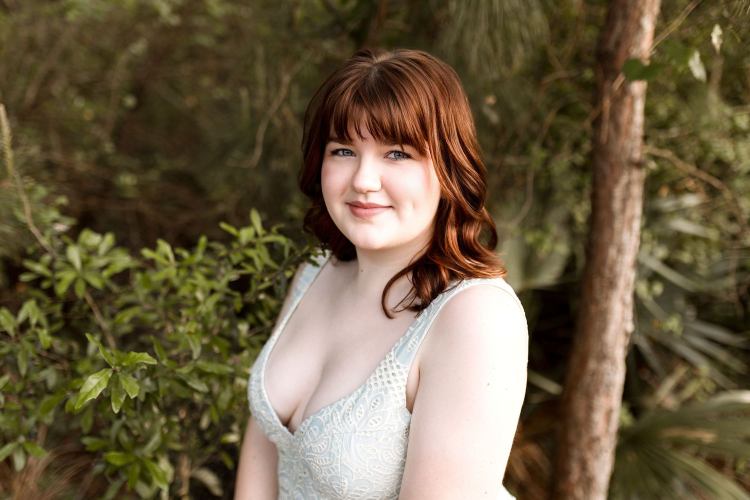 Young woman with red hair in a light green dress smiling outdoors among lush green tropical plants.