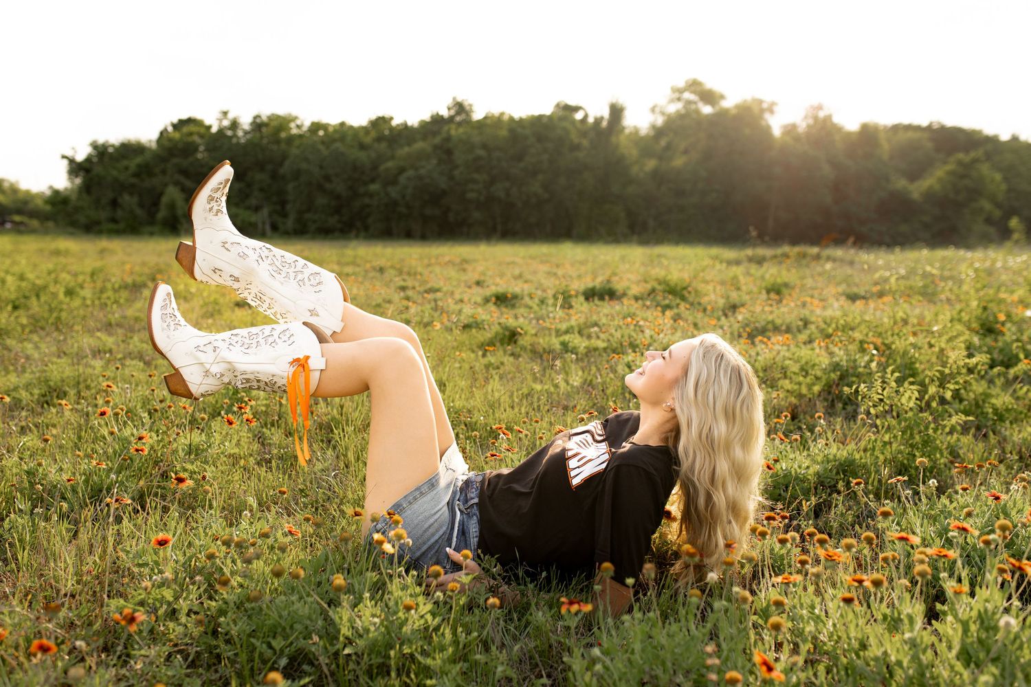 Blonde woman lying in wildflower field wearing denim shorts, black top, and white cowboy boots raised in the air.