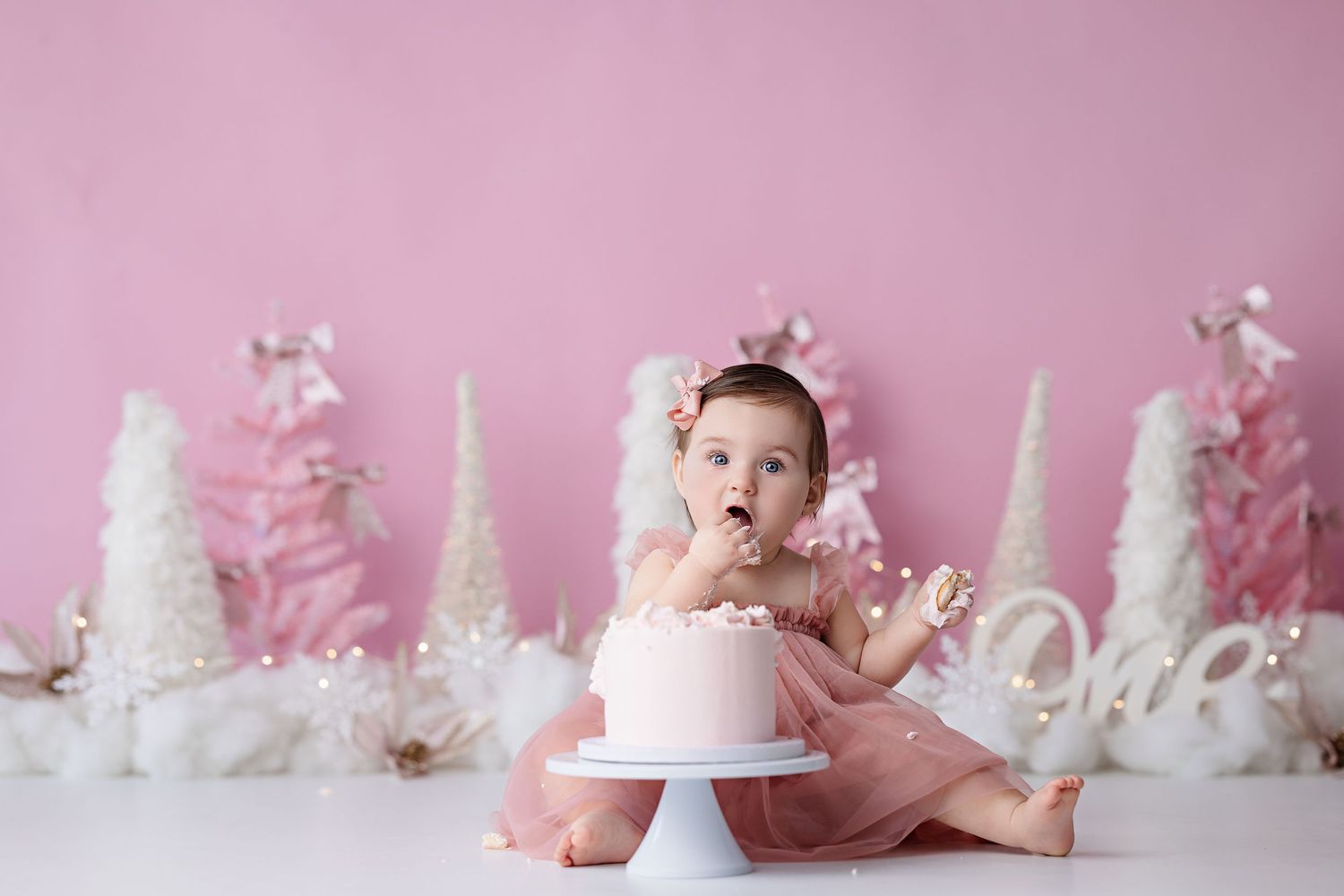 Baby in pink tulle dress sits beside small cake with winter wonderland themed decorations and white trees in background.