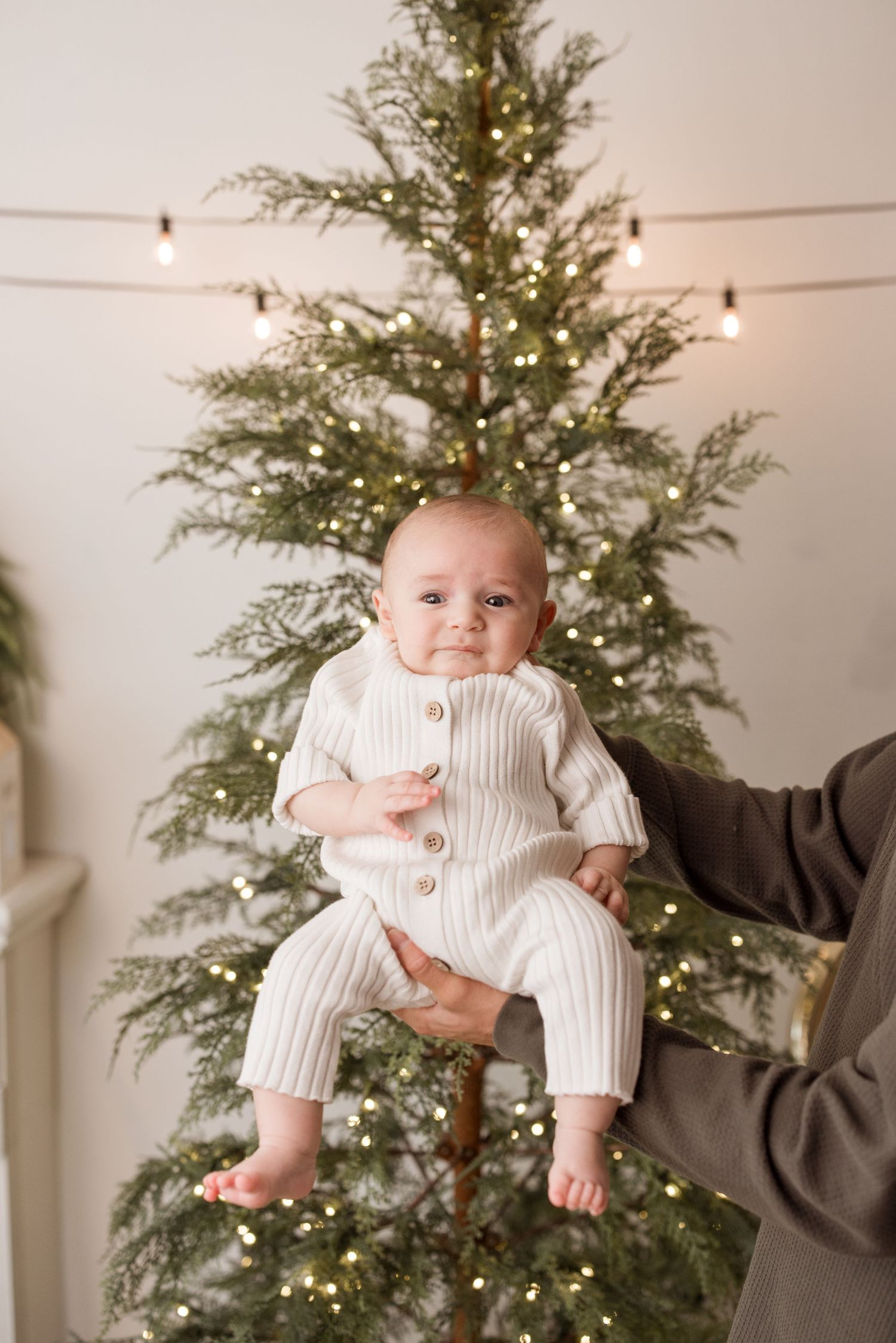 A baby in a white outfit is playfully lifted in front of a decorated Christmas tree with string lights.