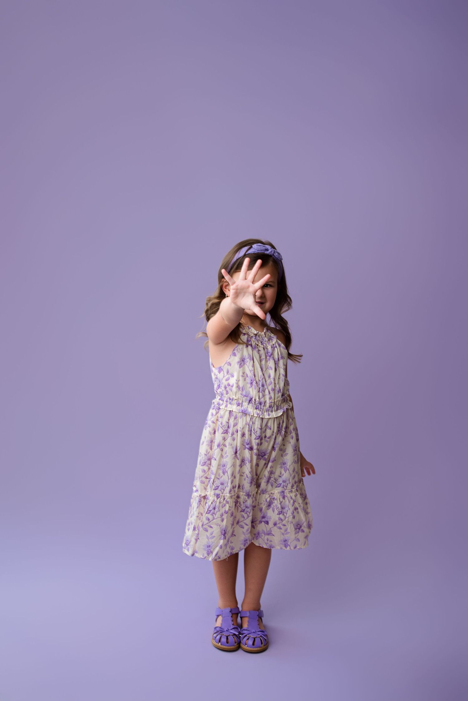 A child in a floral dress with purple sandals stands against a purple background holding their hand up.