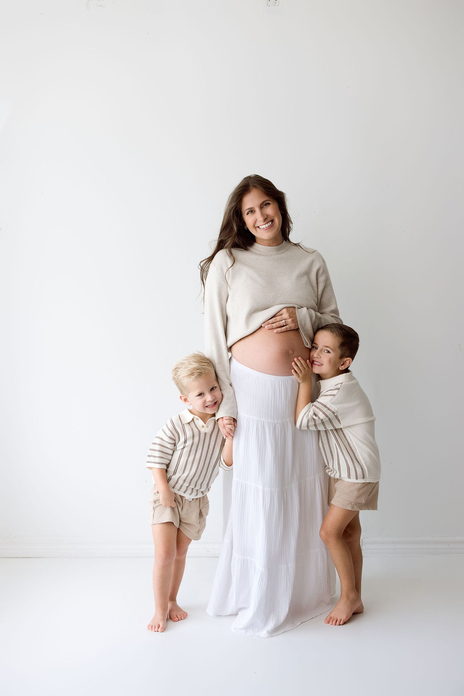 A family wearing white clothing embraces together against a light colored background in a minimalist studio setting.