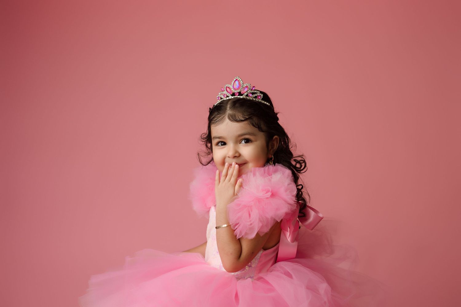 Pink studio portrait with tulle dress and tiara against rosy background.