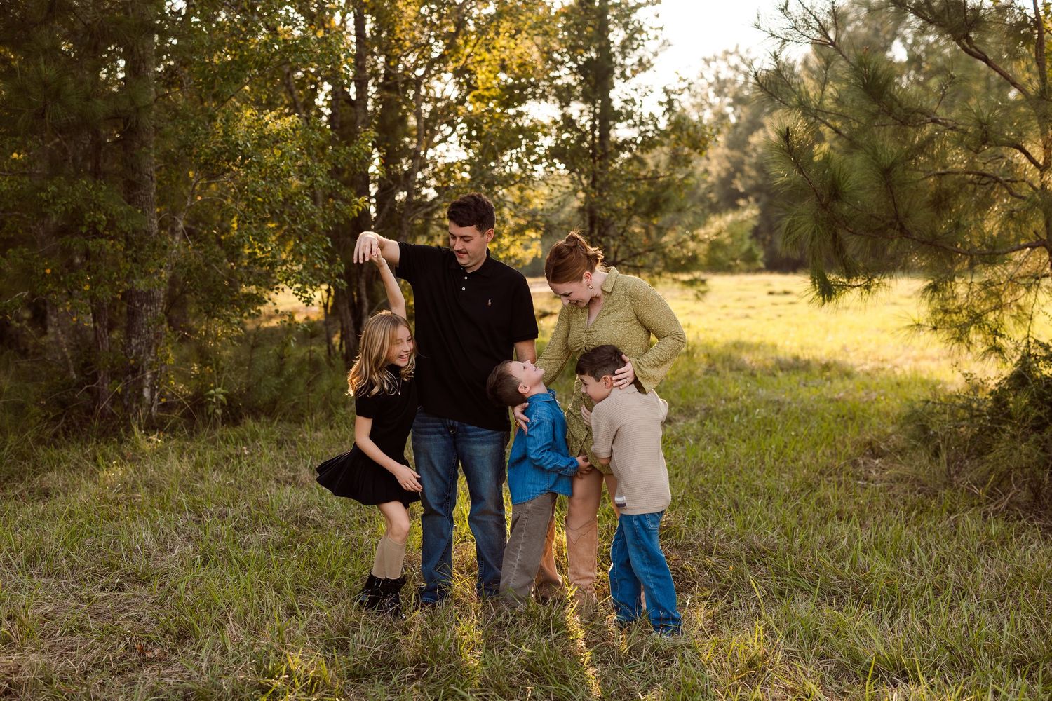 Family of five in a grassy field with tall pine trees, standing close together as the parents hold hands with three children at golden hour.