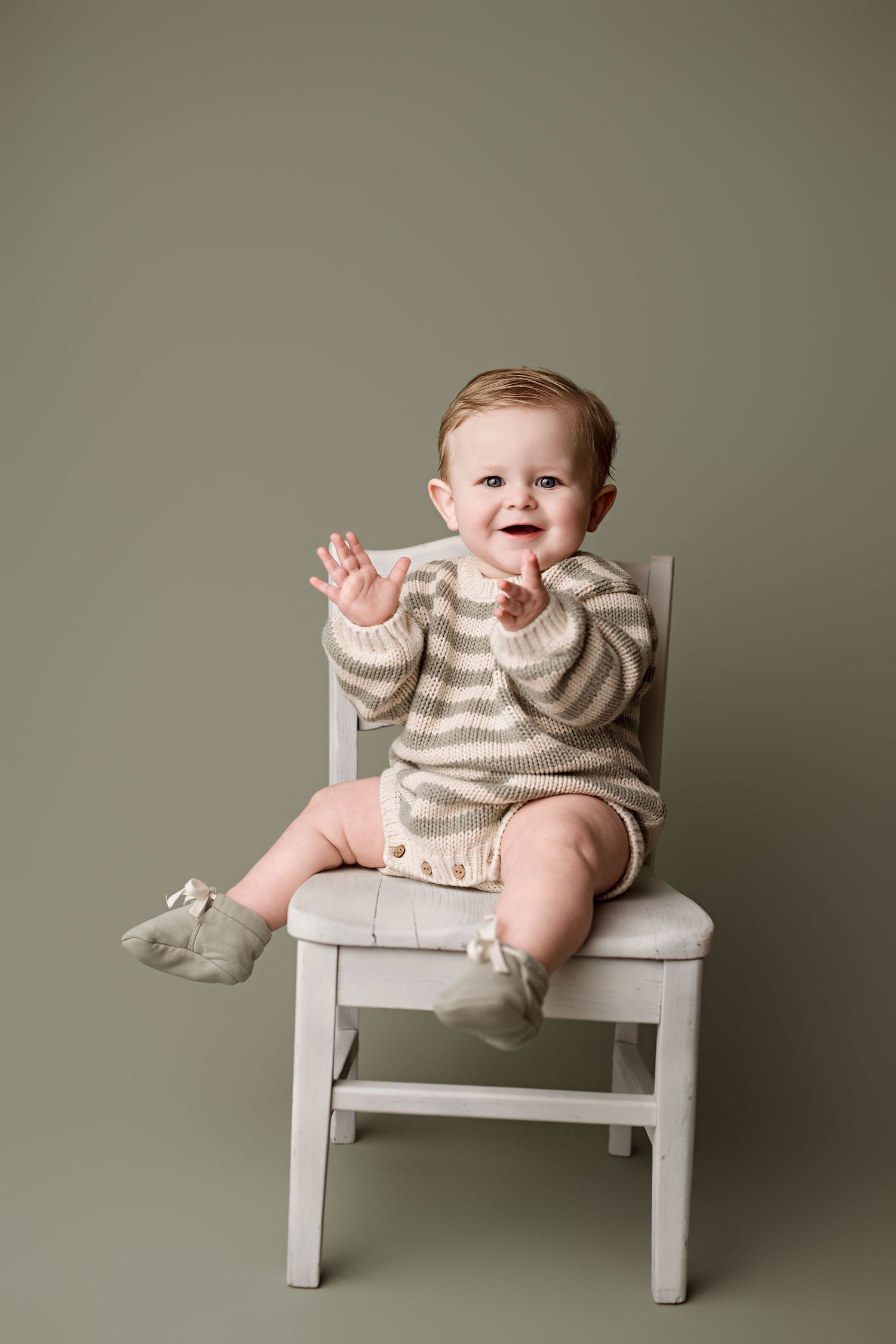 A happy baby in a beige sweater sits and laughs while posing on a small white wooden chair.