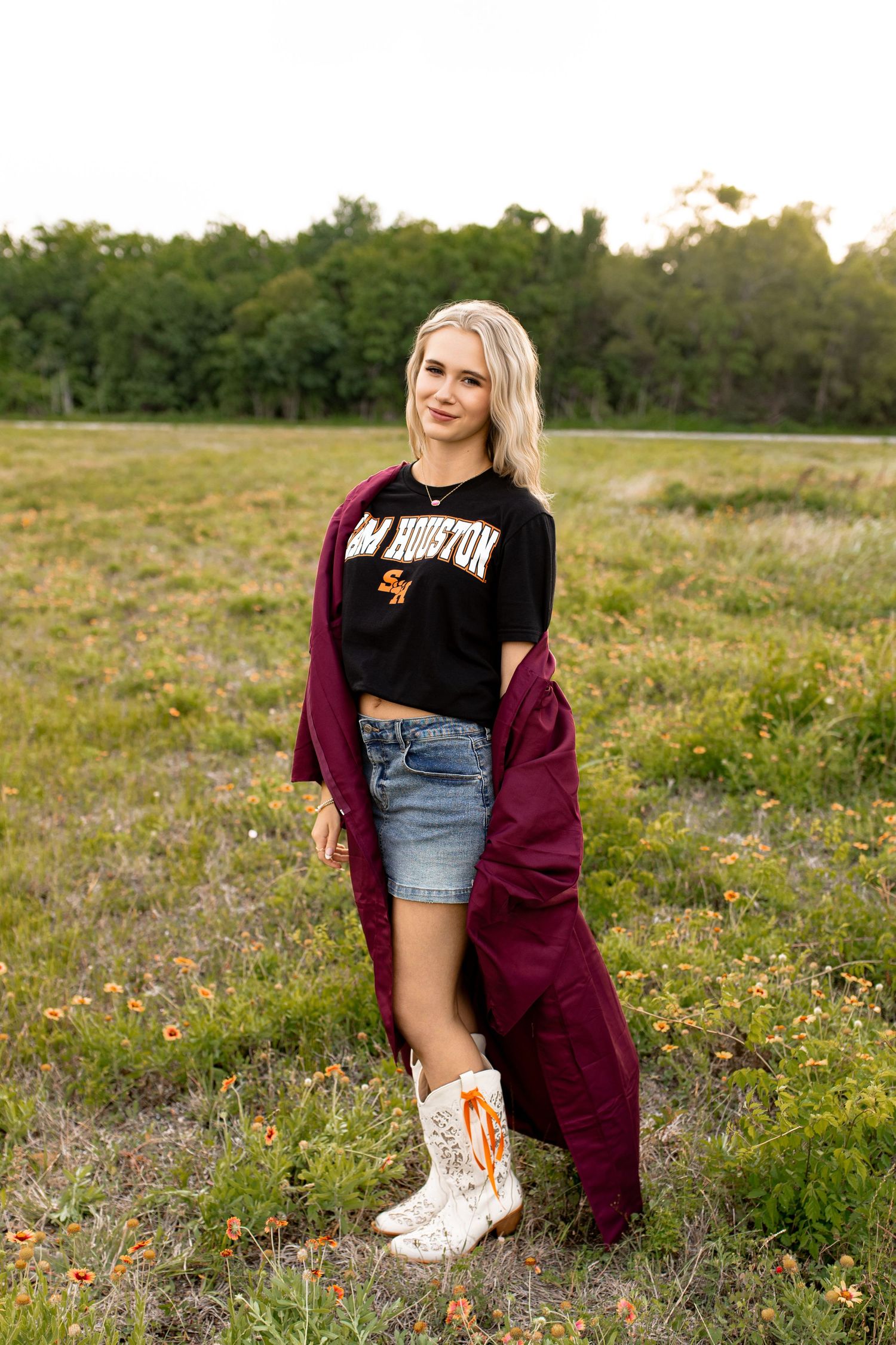 Blonde woman in Sam Houston shirt, denim skirt, and cowboy boots poses in a wildflower field at golden hour. Bree Elle Seniors Beaumont, TX